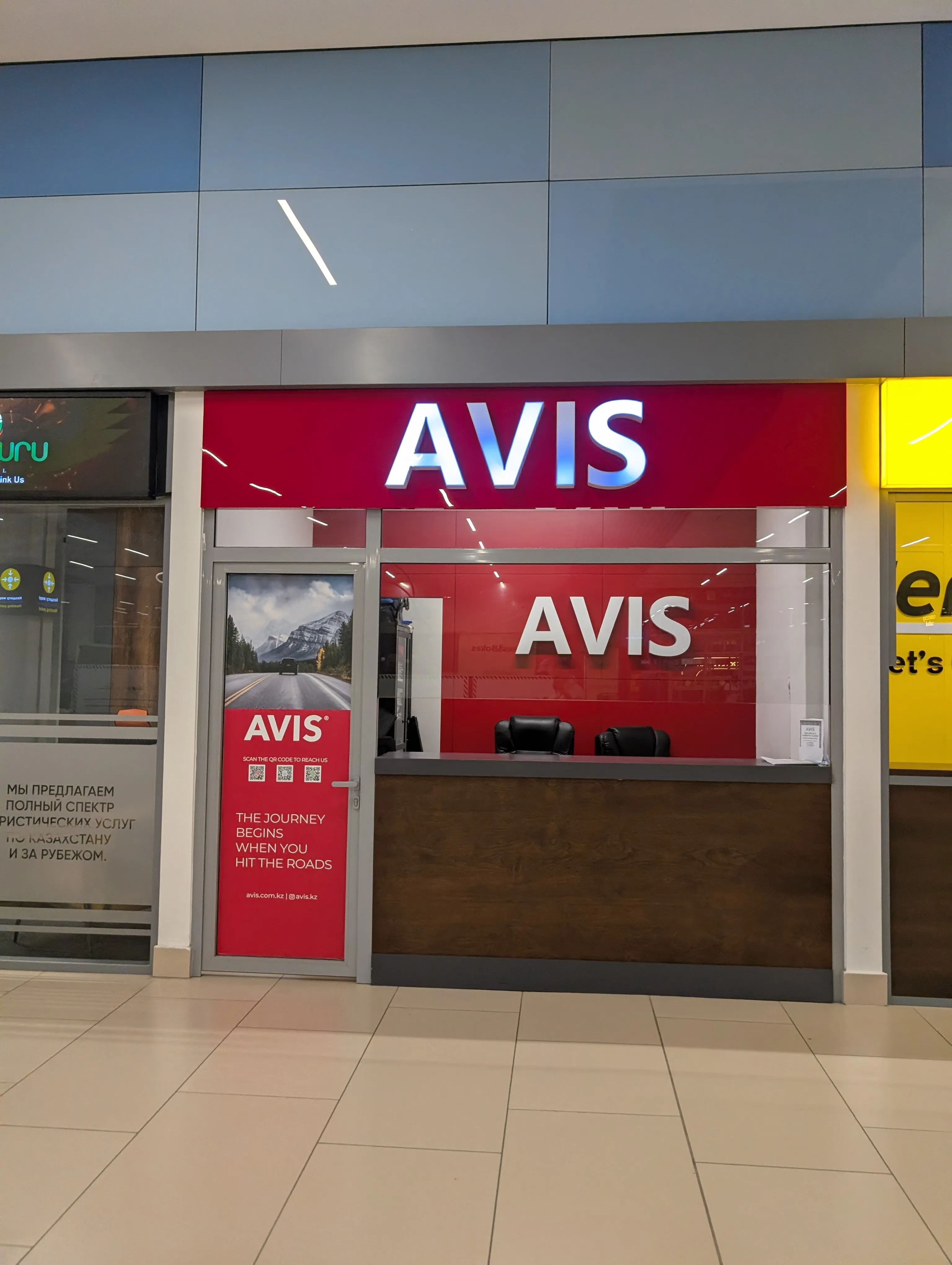 The Avis counter in the arrivals hall at Almaty airport: a lit red fascia over a glass front, a wooden desk with two office chairs behind it, and a poster on the door reading 'the journey begins when you hit the roads'
