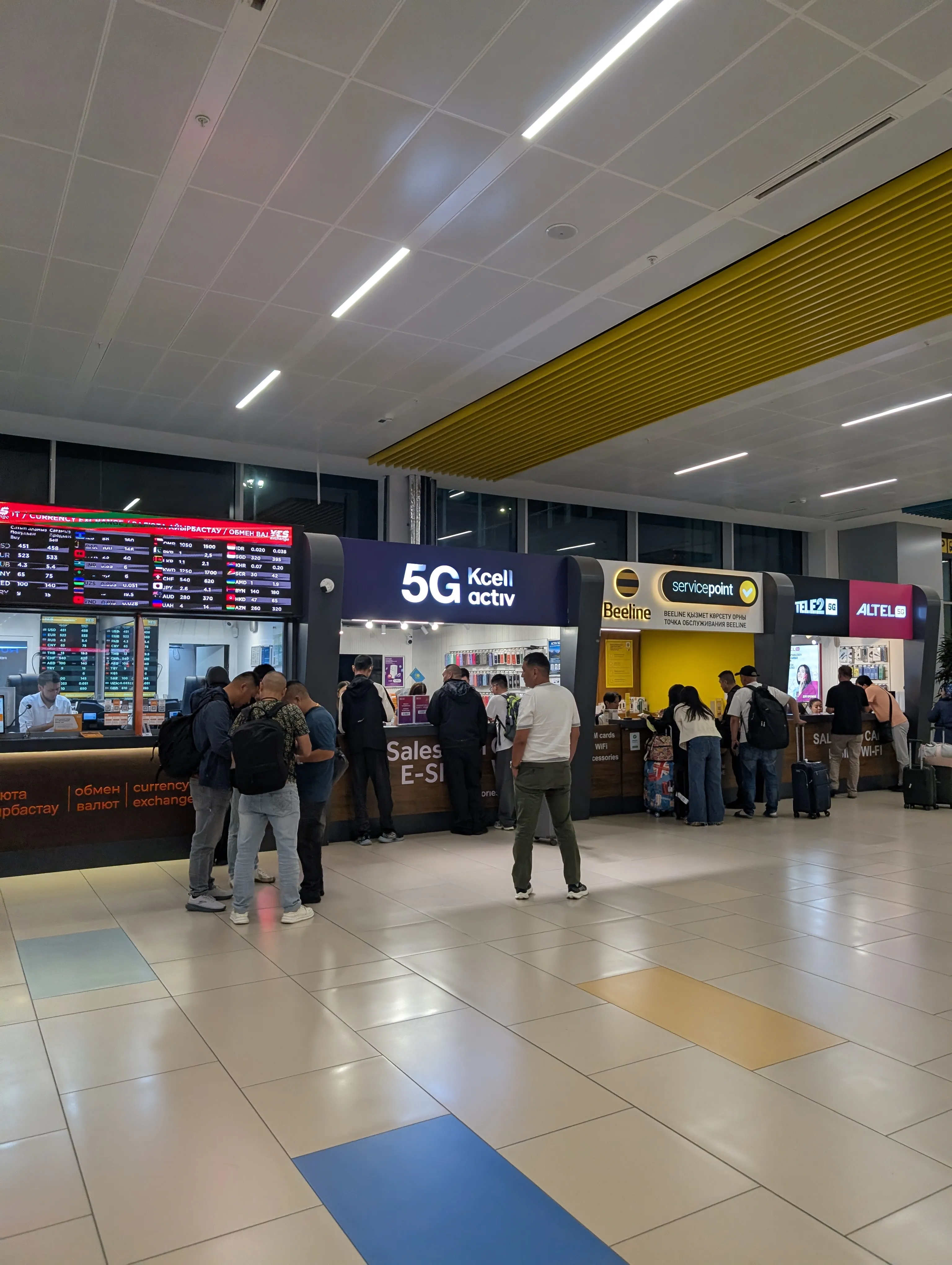 The row of operator counters in the Almaty arrivals hall: a currency exchange board with rates, a counter lettered '5G Kcell activ' and 'sales e-SIM', then Beeline, Tele2 and Altel, with arriving passengers queueing at each of them alongside their luggage