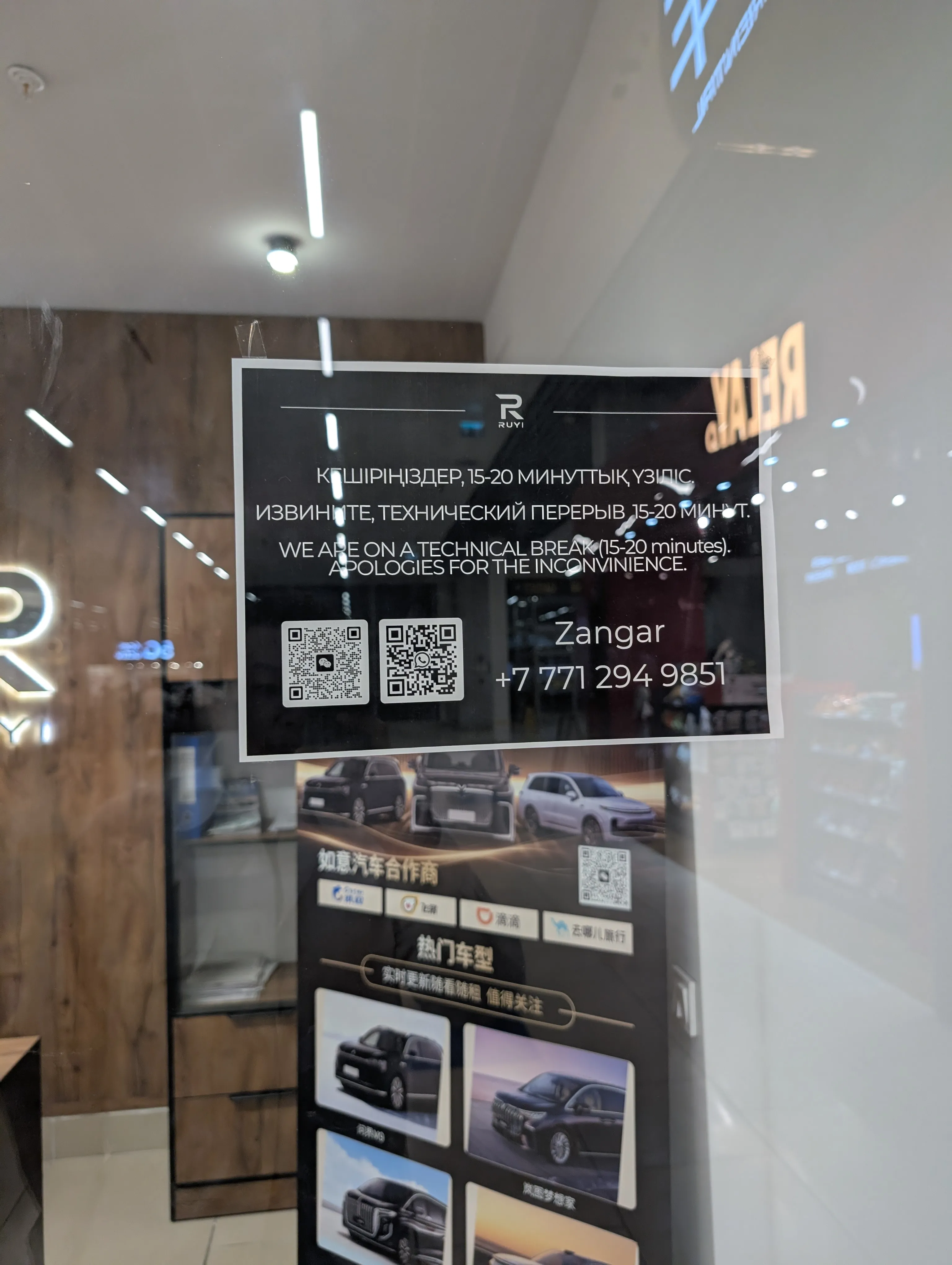 A notice taped inside the glass at the Ruyi counter, printed in Kazakh, Russian and English: a technical break of fifteen to twenty minutes, with two QR codes, a name and a phone number under them, and a stand of car models behind the glass