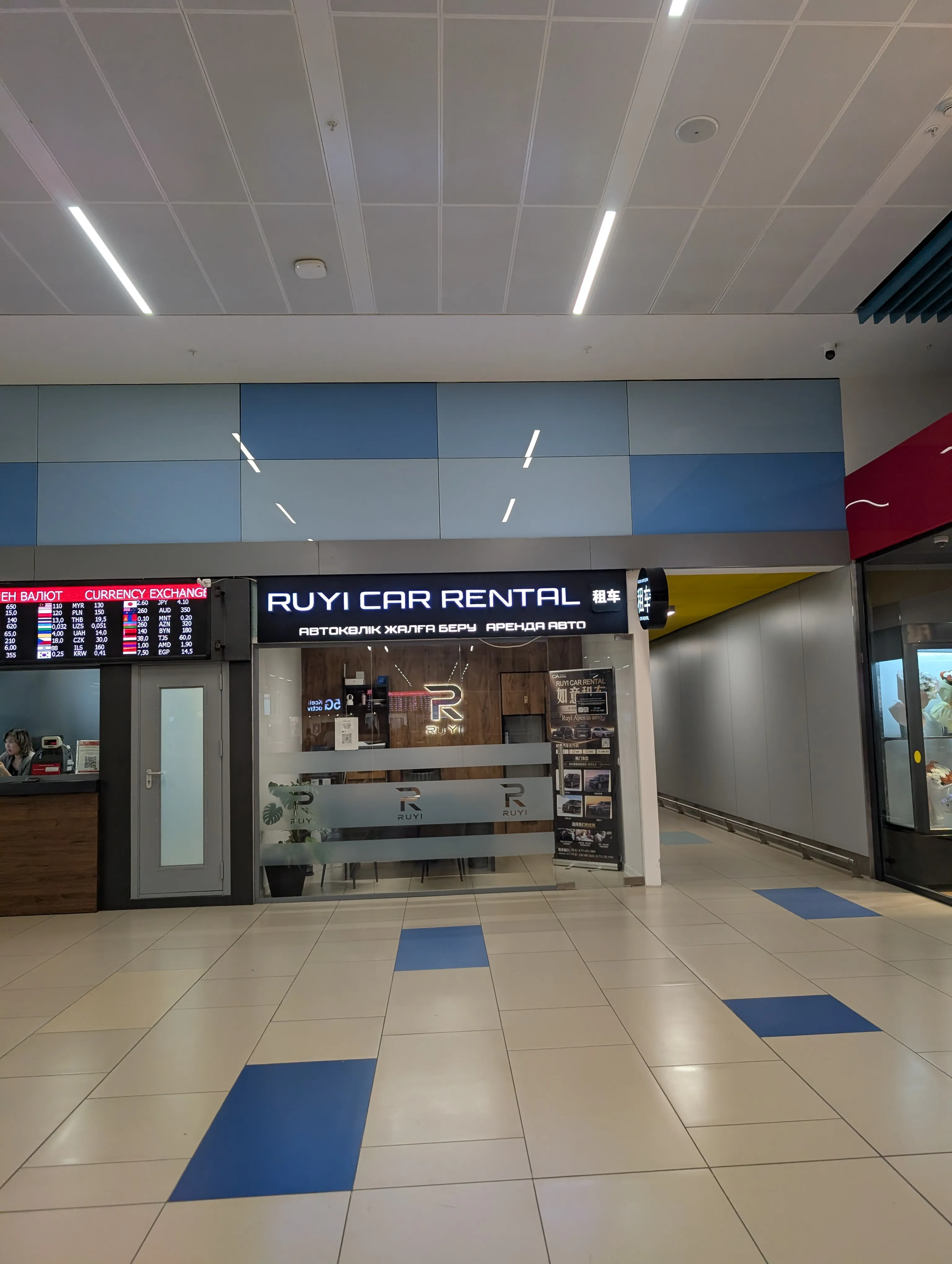 The Ruyi Car Rental counter at Almaty airport: a black fascia lettered in Latin, Kazakh, Russian and Chinese, a lit logo on the wall behind the empty desk, and a currency exchange board carrying rates to the left of it