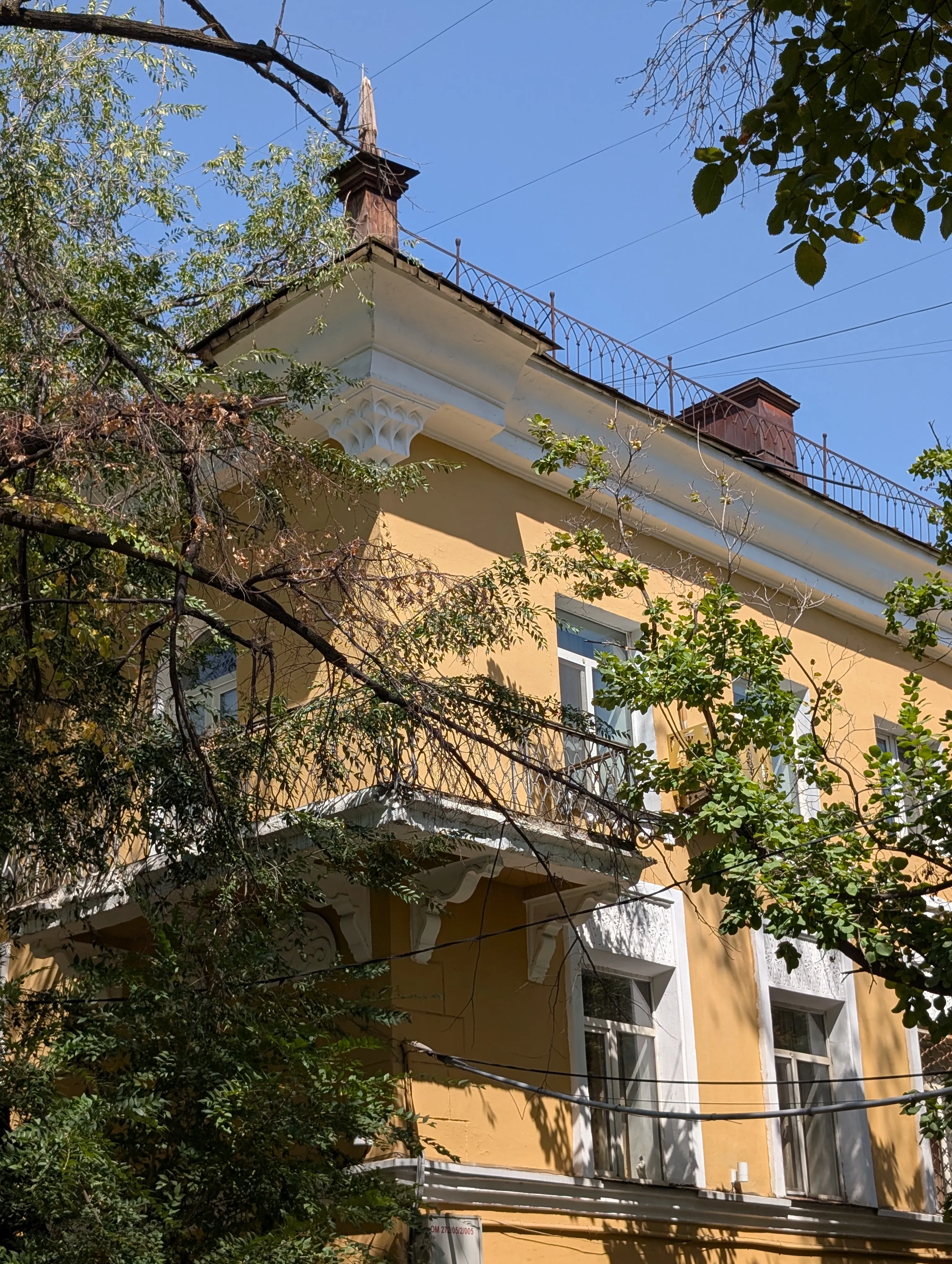 The upper corner of an ochre building: white plaster brackets under the eaves, a decorative wrought-iron railing running along the roofline, and a small wooden finial rising above it