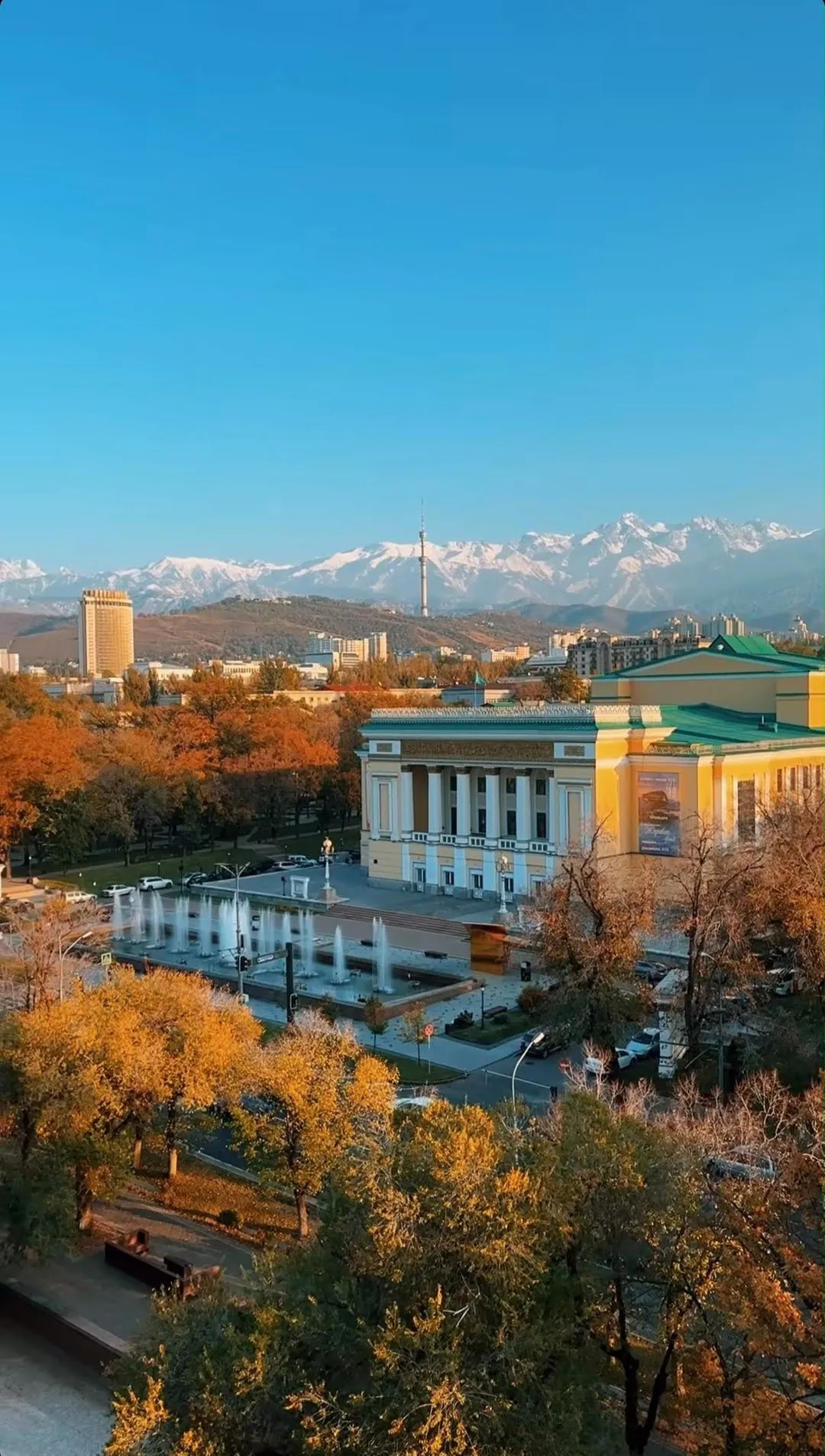 Almaty from above in autumn: the columned opera house and its fountains among orange trees, the Hotel Kazakhstan standing off to the left, the television tower on the ridge, and the snow line of the Tien Shan across the whole horizon