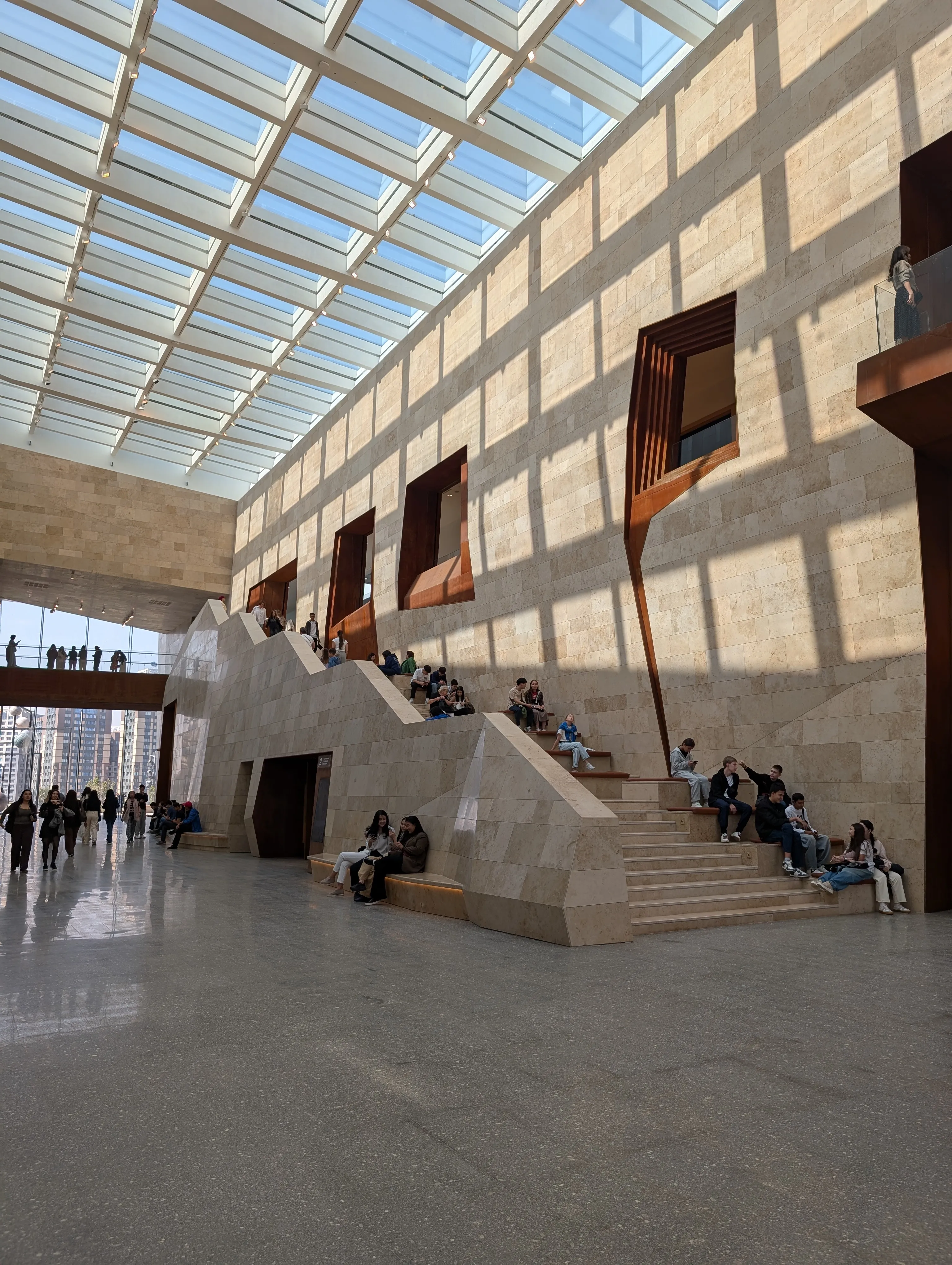 The main hall of the Almaty Museum of Arts: pale travertine walls rising to a glazed roof, a broad stone staircase with people sitting on the steps, and openings lined in rust-coloured steel