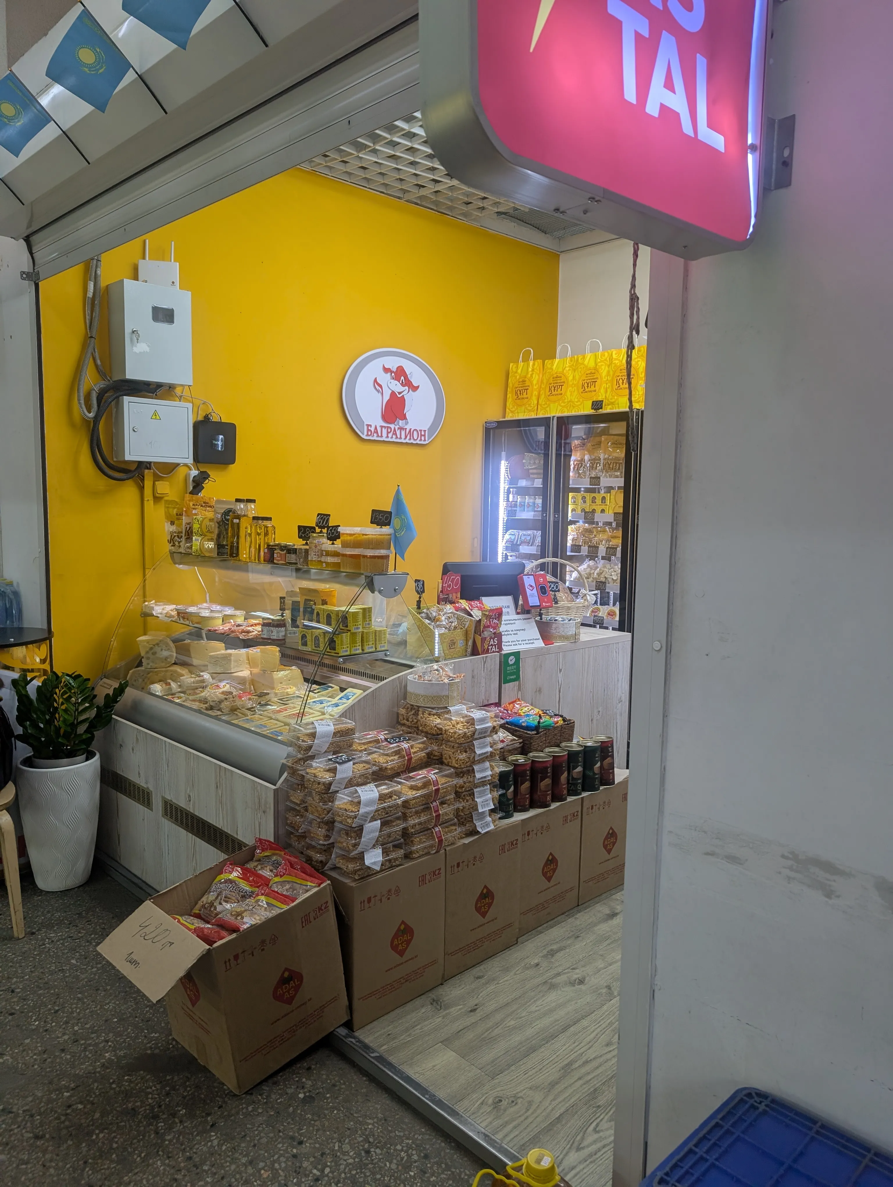 The Bagration counter: a red cow logo on a yellow wall above a chilled case of cheese wheels, jars and tubs of honey along the top shelf, a small Kazakh flag by the till, and stacked boxes of chak-chak with price tickets beside an open carton of baursak marked 420 tenge