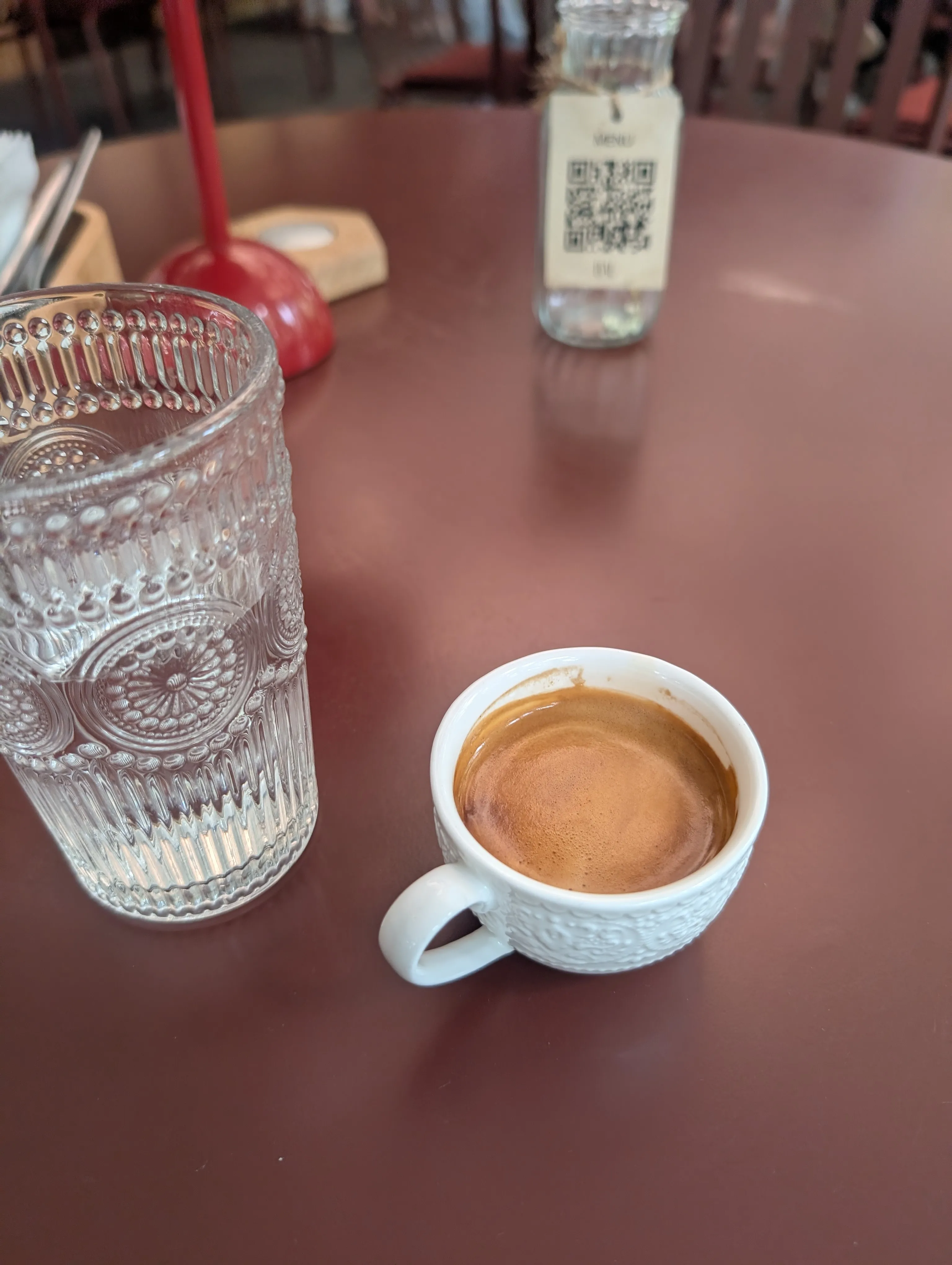 Espresso in a white cup on a red table at Bal, with a water glass and the QR menu