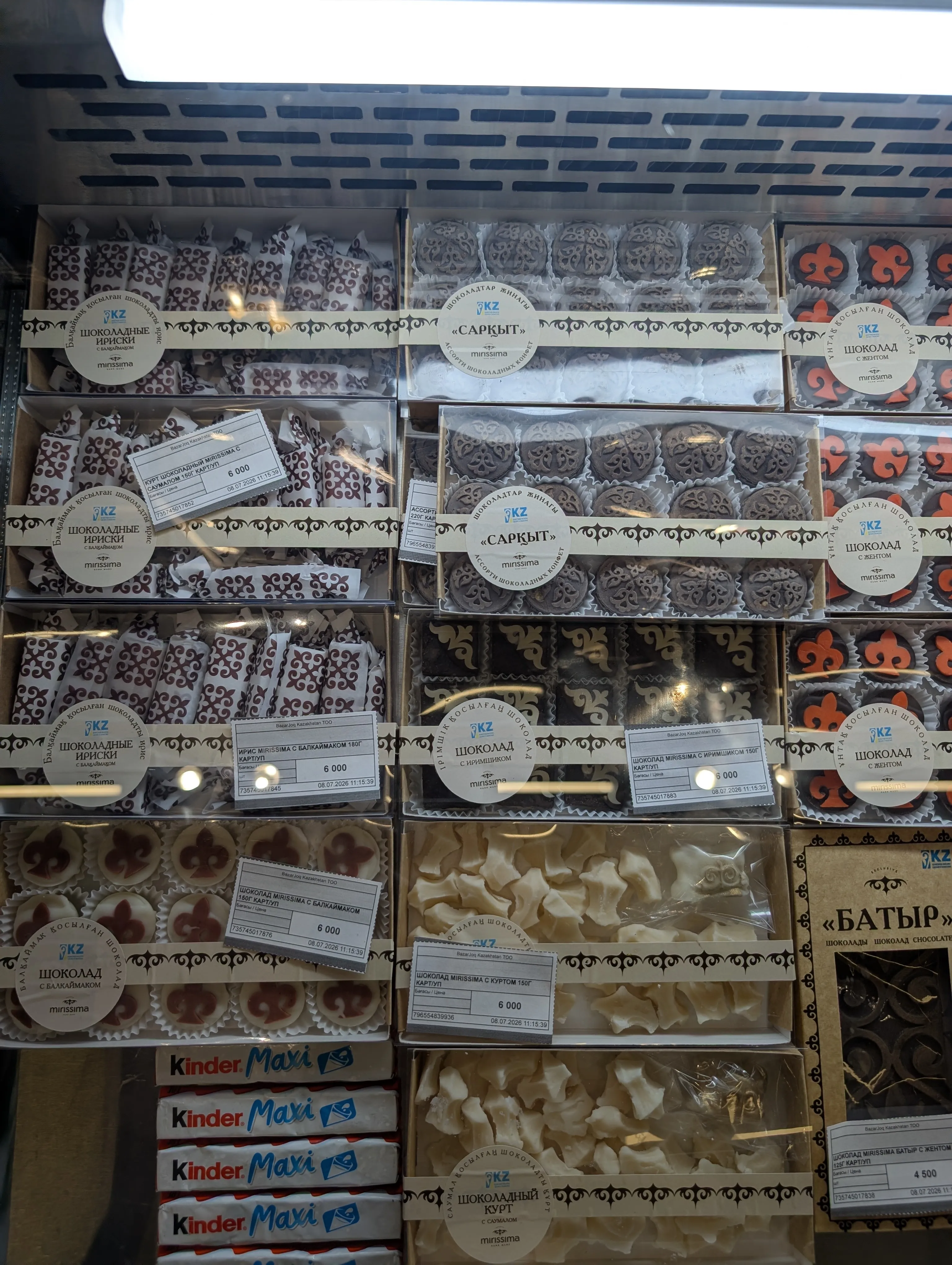 The chilled case at Bazarjoq inside the Green Bazaar: trays of Mirissima chocolate — dark rounds stamped with Kazakh ornament, toffees in patterned wrappers and white chocolate qurt — each with a printed ticket reading 6 000 tenge