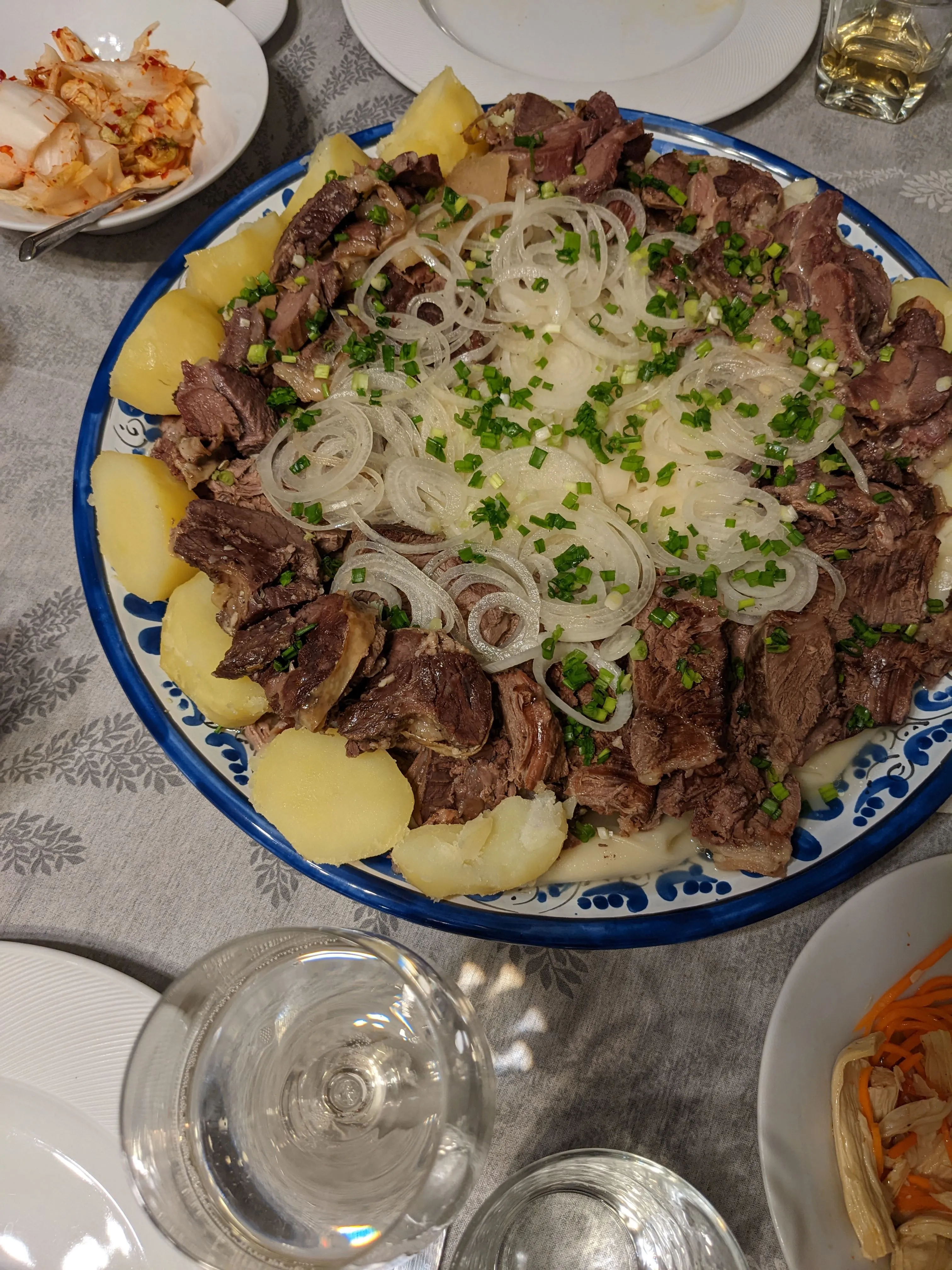 A family-sized beshbarmak at home: a wide platter of boiled meat and potatoes under rings of raw onion