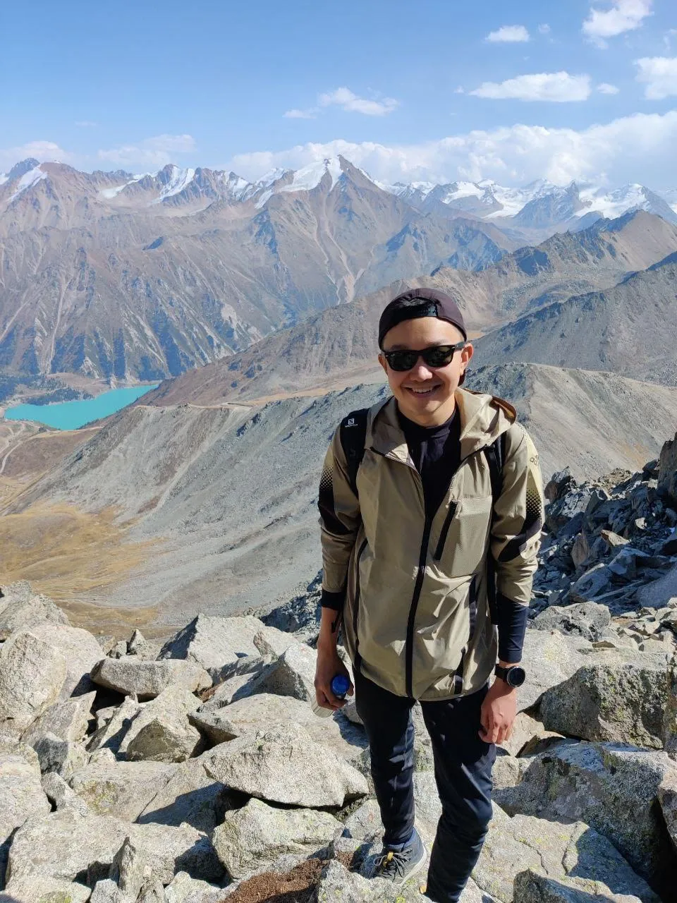 A walker standing on a rocky summit in the range above Almaty, a turquoise glacial lake far below and the snow crest of the Tien Shan filling the horizon behind