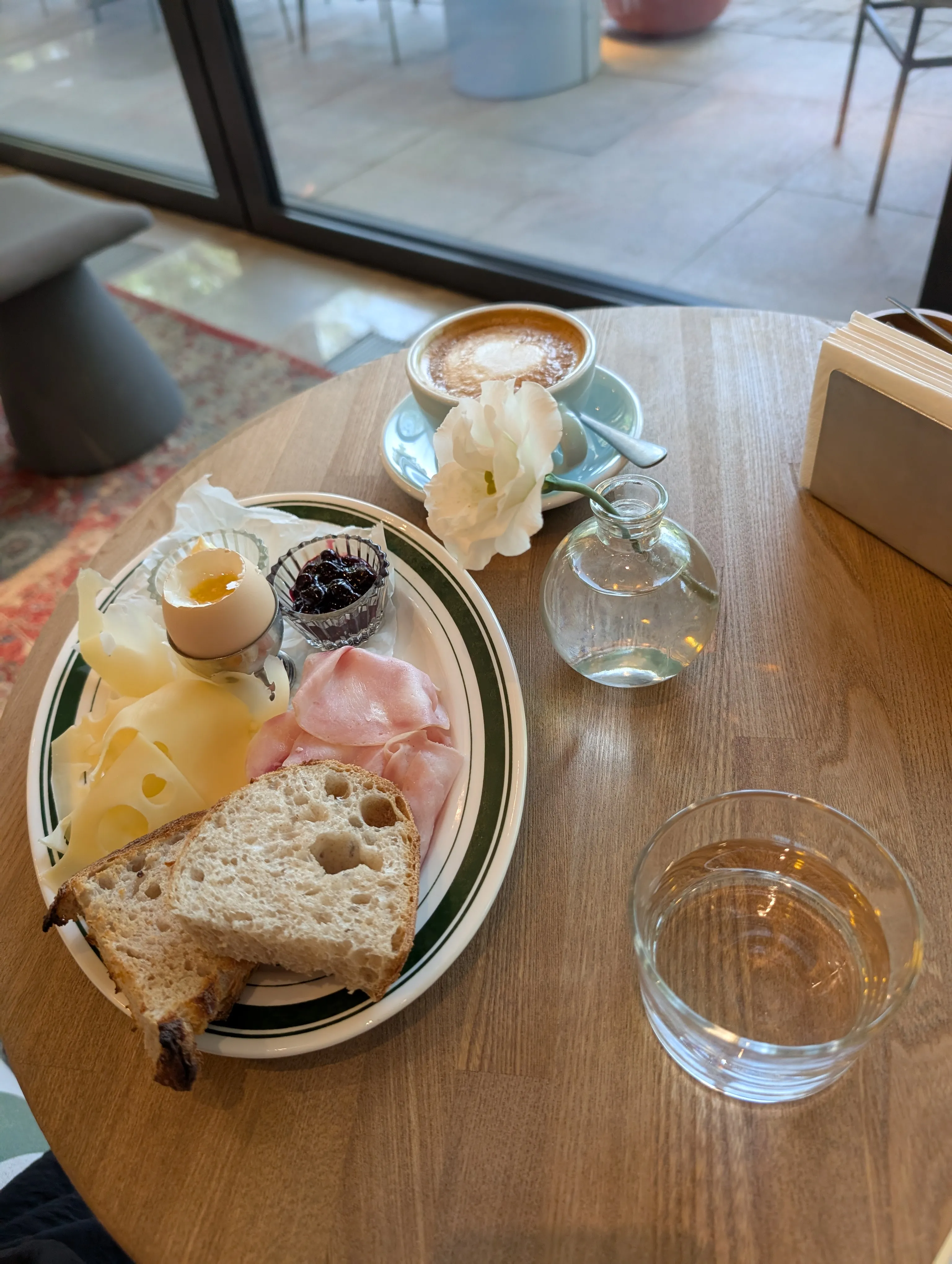 A lighter breakfast at Akku: a soft egg, sliced cheese and ham, sourdough and jam, a cappuccino and a single flower