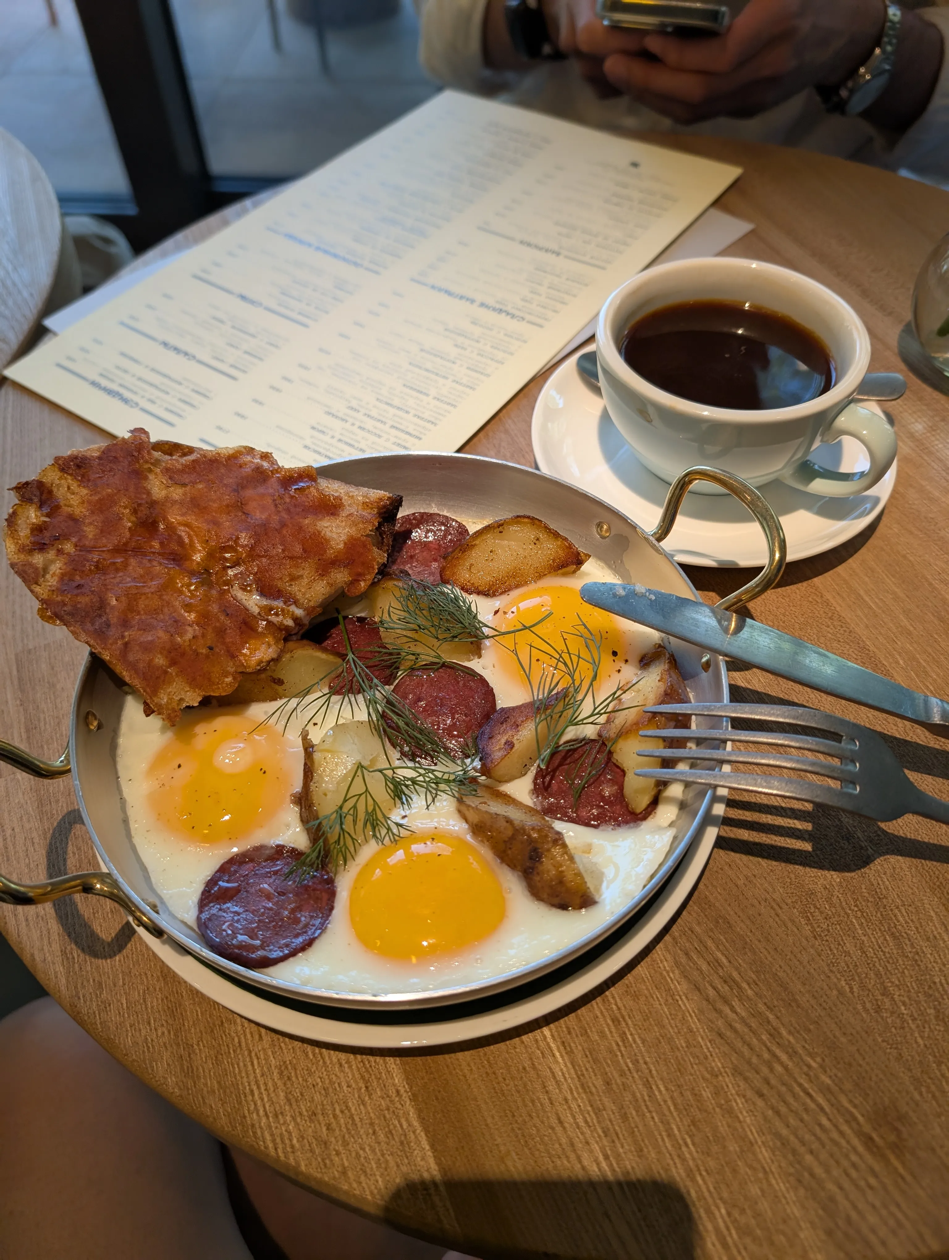Breakfast at Akku: fried eggs, sujuk, a hash brown and dill on a steel plate, with a cup of coffee and the paper menu beside it