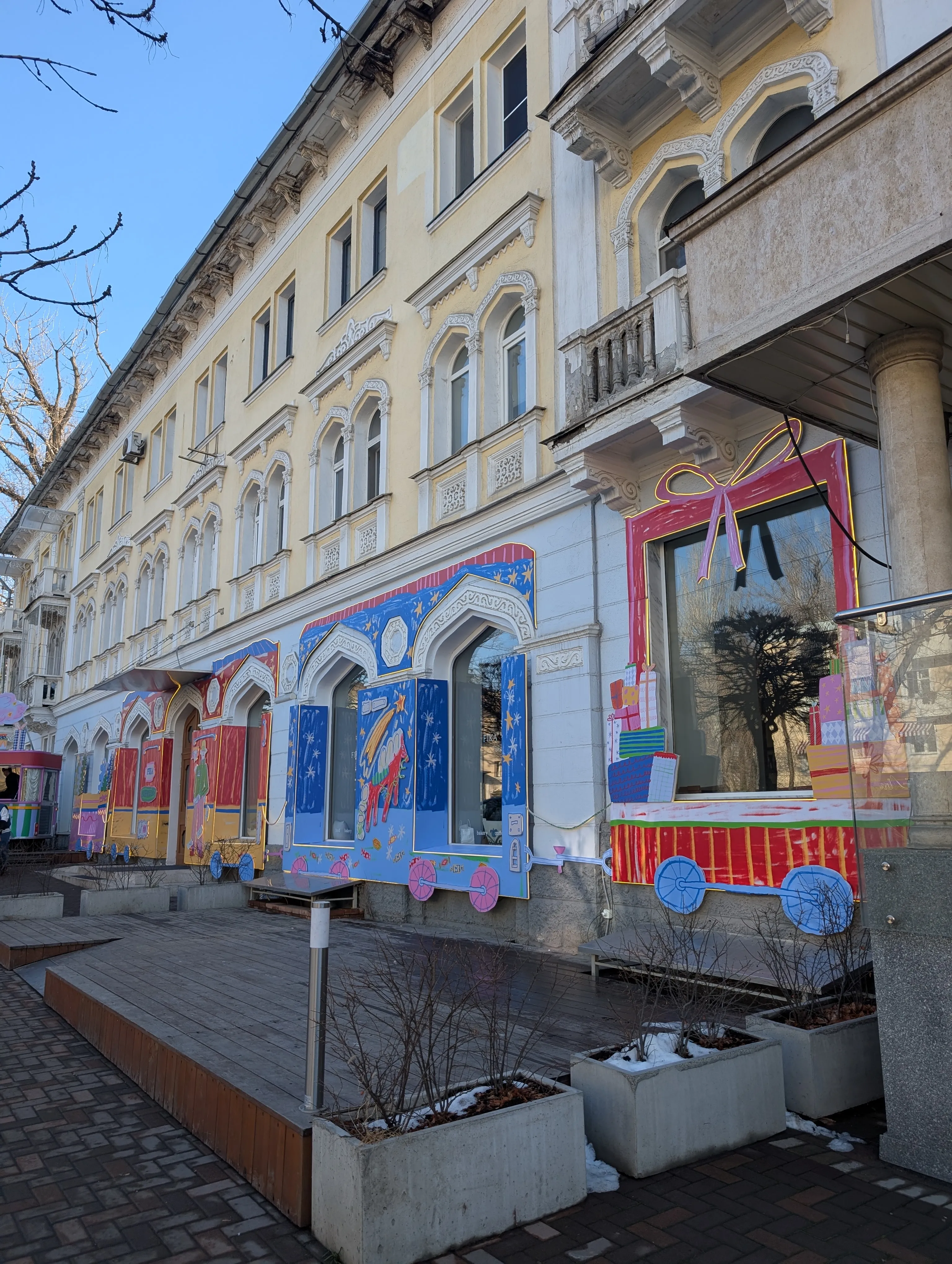 The Turksib House at Kabanbai Batyr 104, the building Fika is in, dressed for the new year: painted panels of a festive train and stacked presents across the arched windows, a pink bow over the corner glass, and snow in the planters below