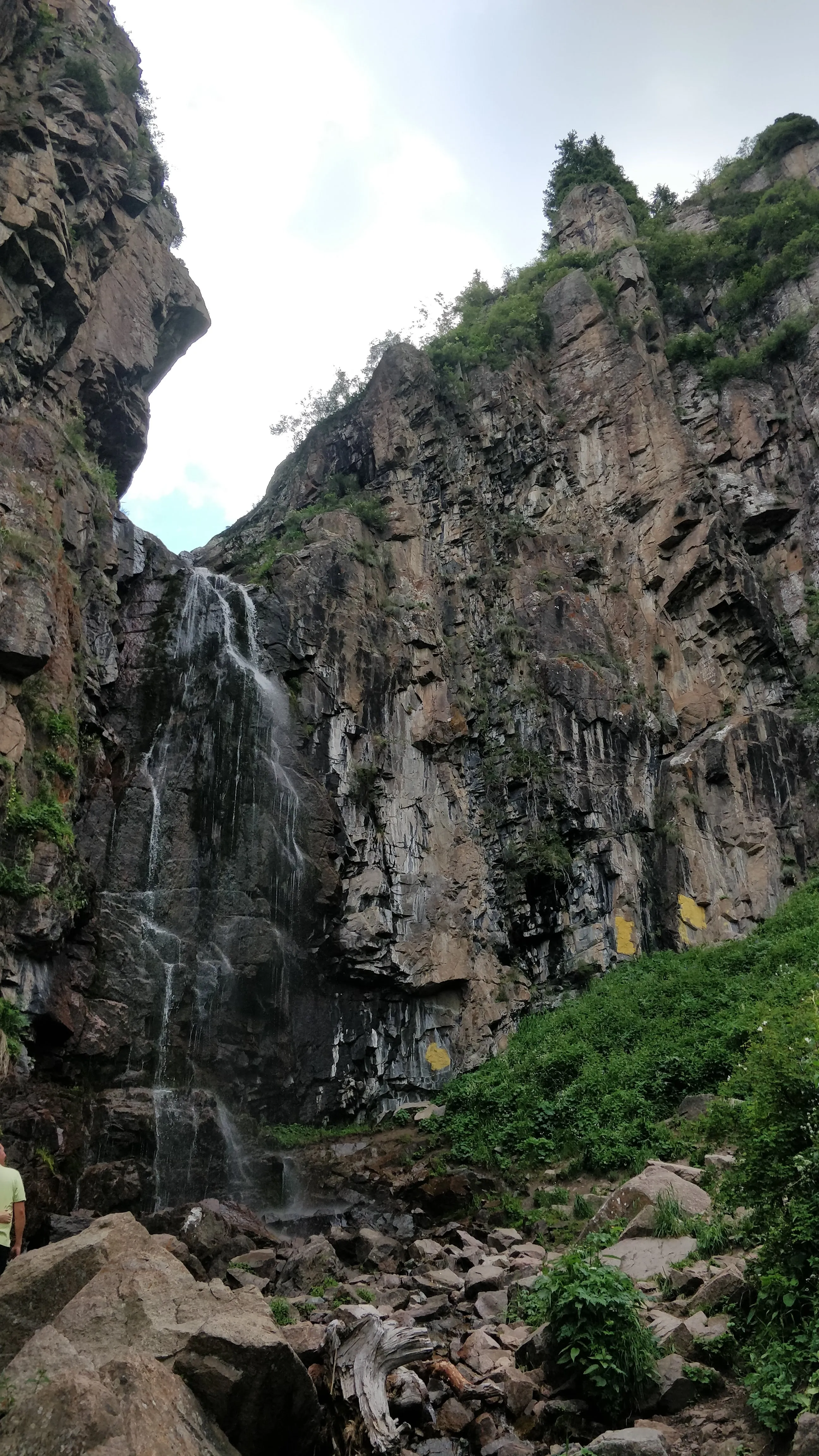 The waterfall in the Butakovka gorge: a thin fall dropping down a dark rock face streaked with water, green slopes and scree at its foot, and a walker standing at the bottom for scale