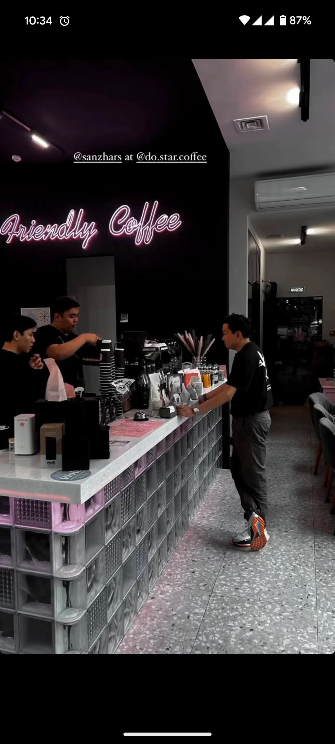 The counter at Do.Star under its pink neon script reading Friendly Coffee: two baristas working the espresso machine behind a terrazzo bar whose front is stacked with pink and clear crates, and a customer waiting at the far end