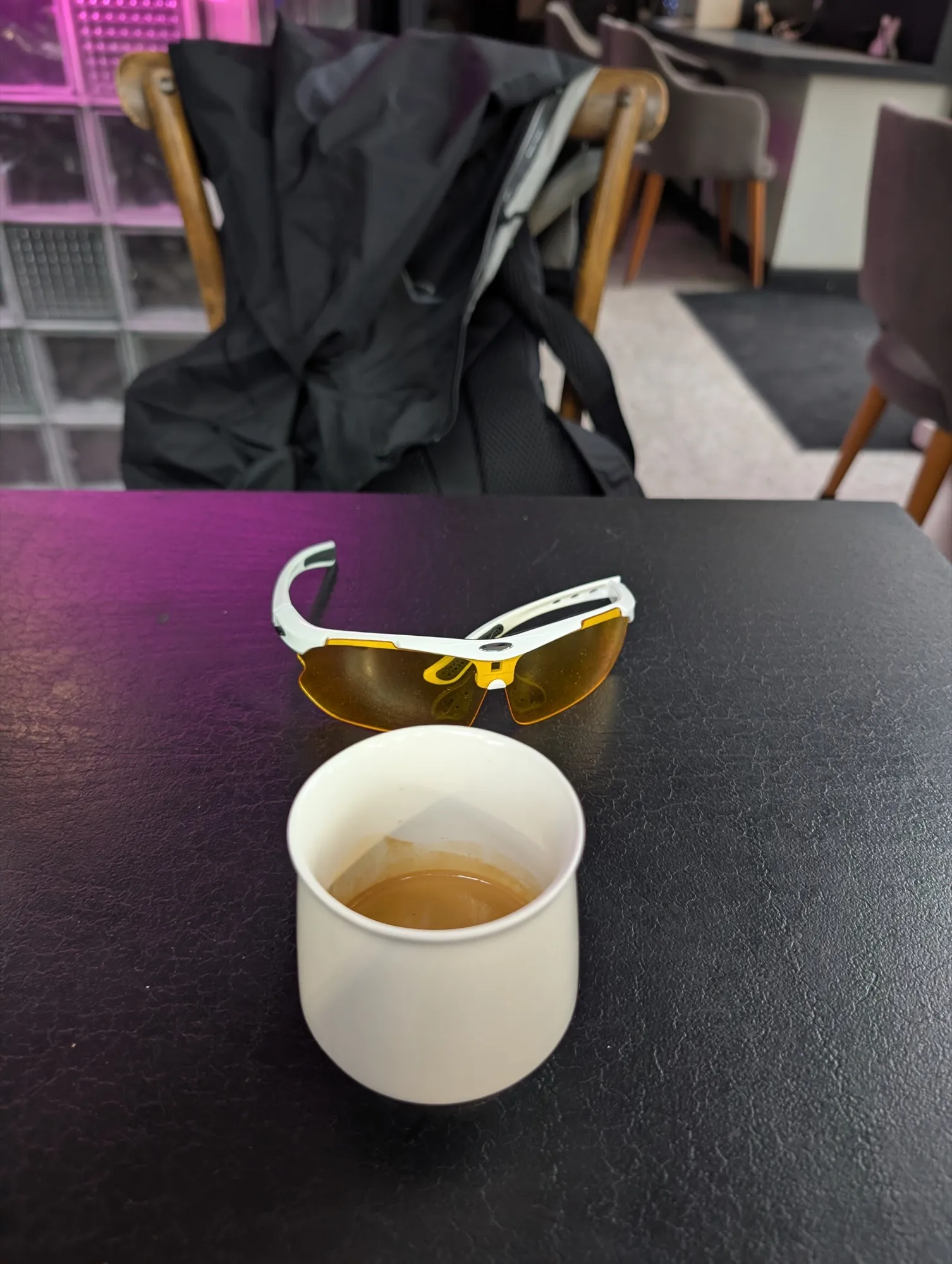 An espresso in a white cup on the black table at Do.Star, a pair of white and yellow sports sunglasses lying beside it and a jacket over the wooden chair behind, with the cafe's pink glass-block wall out of focus beyond