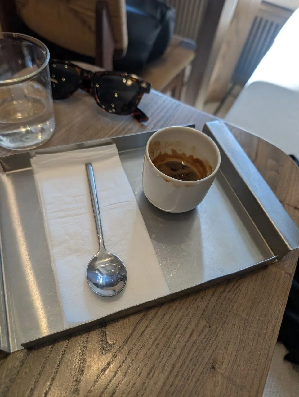 A finished espresso at Julius on Kabanbai Batyr: the white cup with crema traces stands on a steel tray beside a paper napkin and spoon, with a glass of water and a pair of tortoiseshell sunglasses on the wooden table