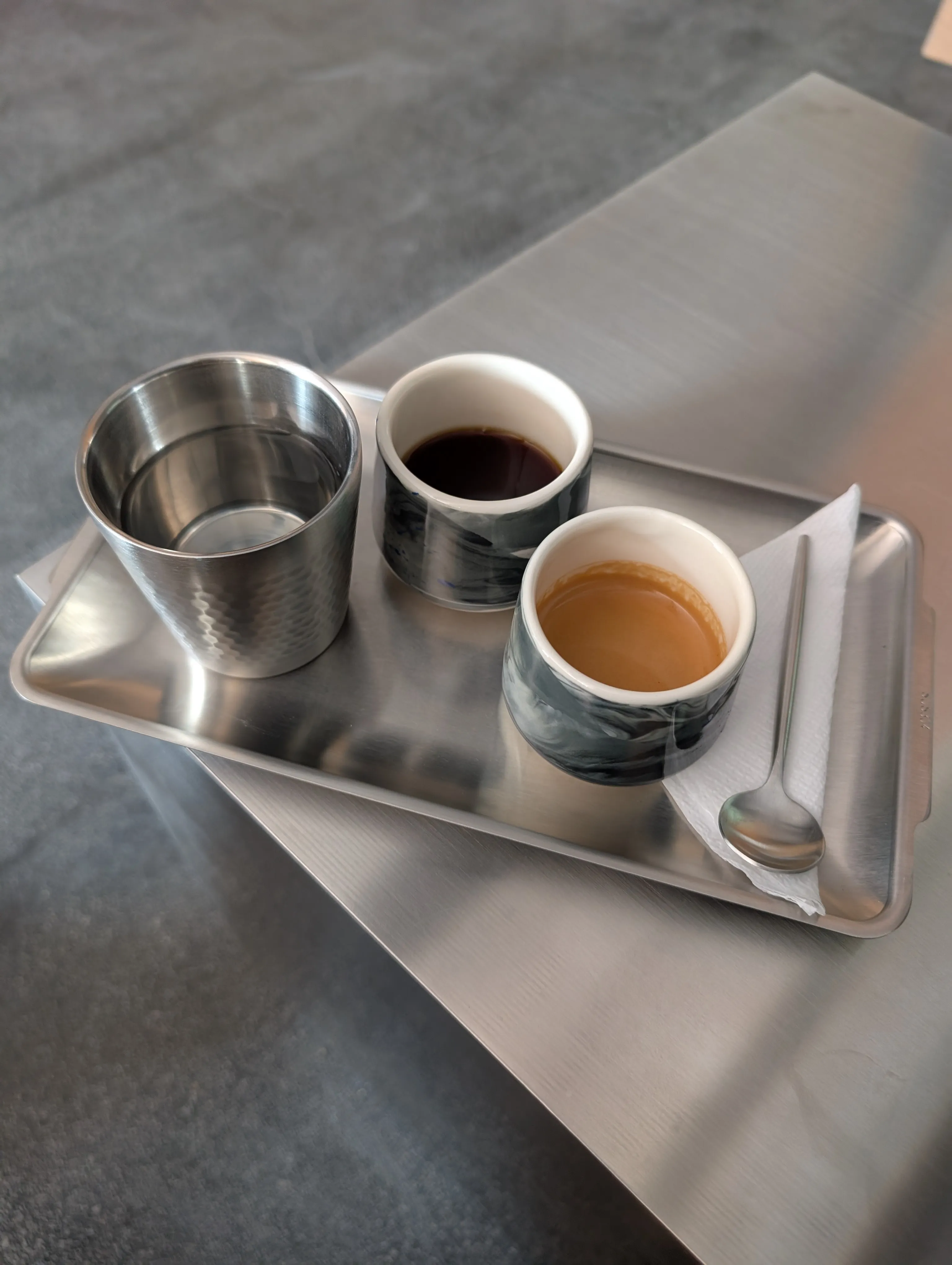 A steel tray at Solid: a hammered steel tumbler of water, a marbled black-and-white cup of dark coffee, and an espresso under its crema in a second marbled cup, with a spoon on a folded napkin