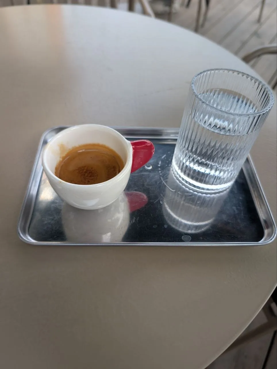 An espresso in a white cup with a red handle at Collab, on a steel tray beside a ribbed glass of water, on a round beige table on the terrace