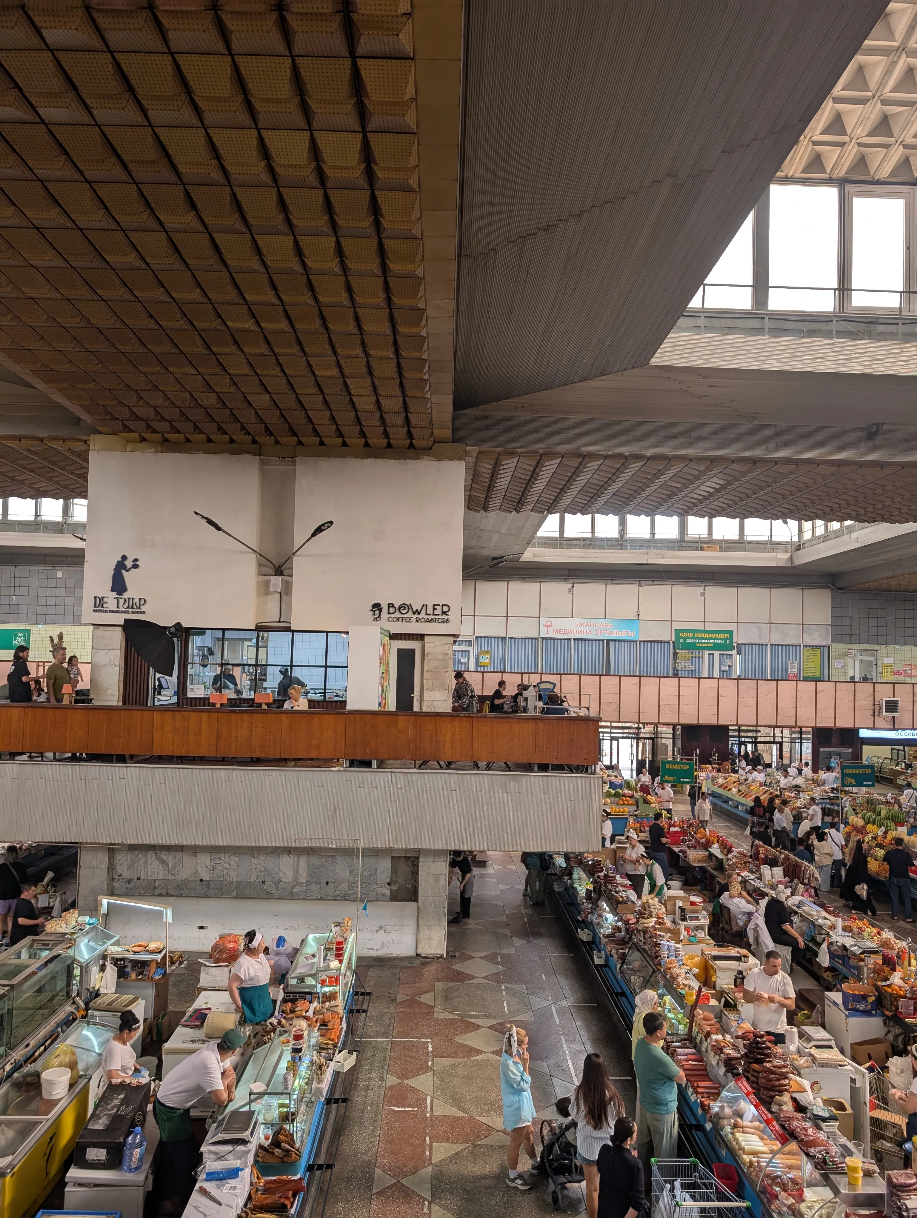 The interior of the Green Bazaar seen from a balcony: long parallel rows of produce stalls filling a wide hall under a single ribbed concrete roof, with no internal columns