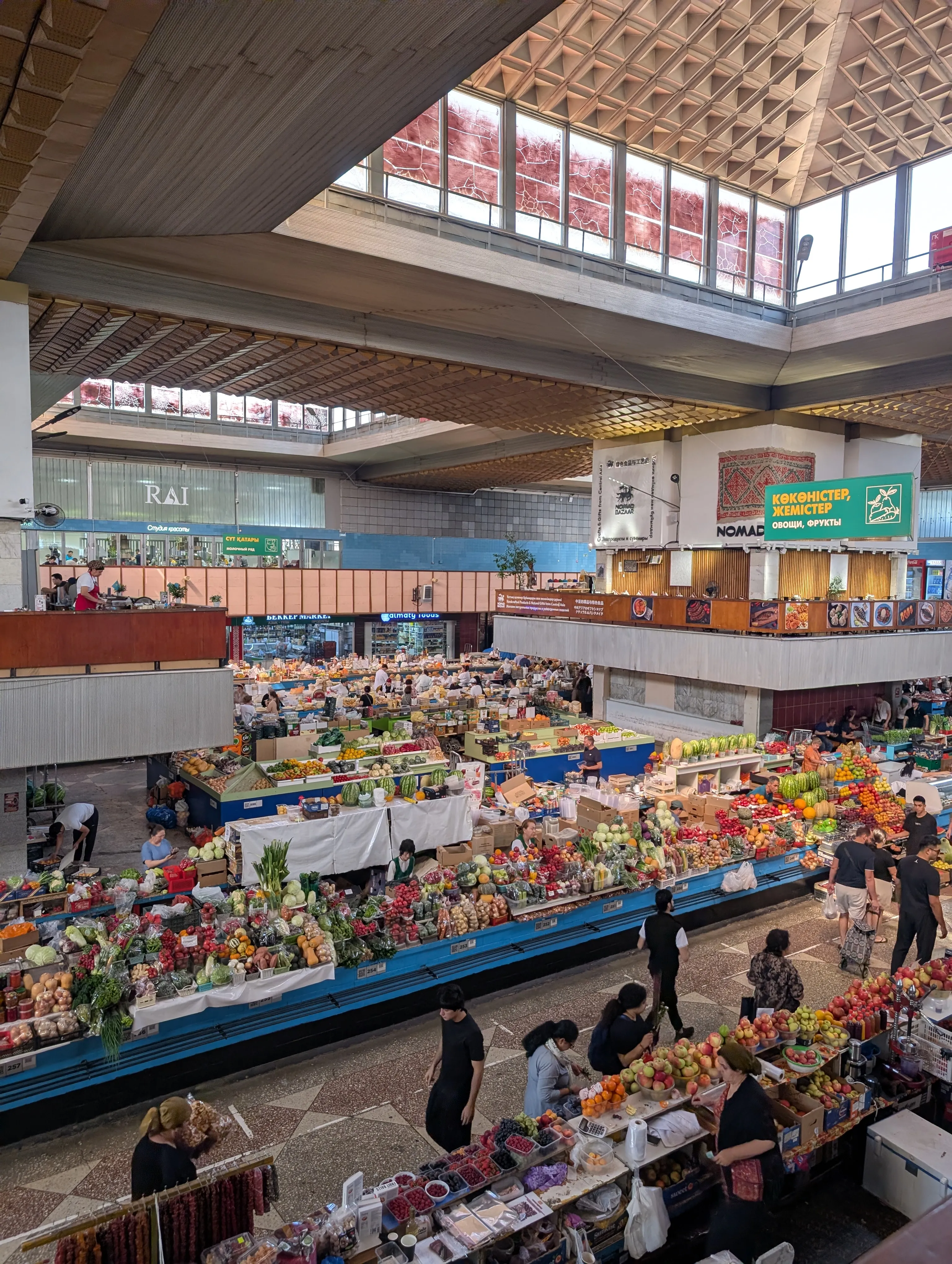 The Green Bazaar from the upper gallery: parallel rows of fruit and vegetable stalls filling the hall below, traders behind them, under a folded concrete roof lit by a band of glazing