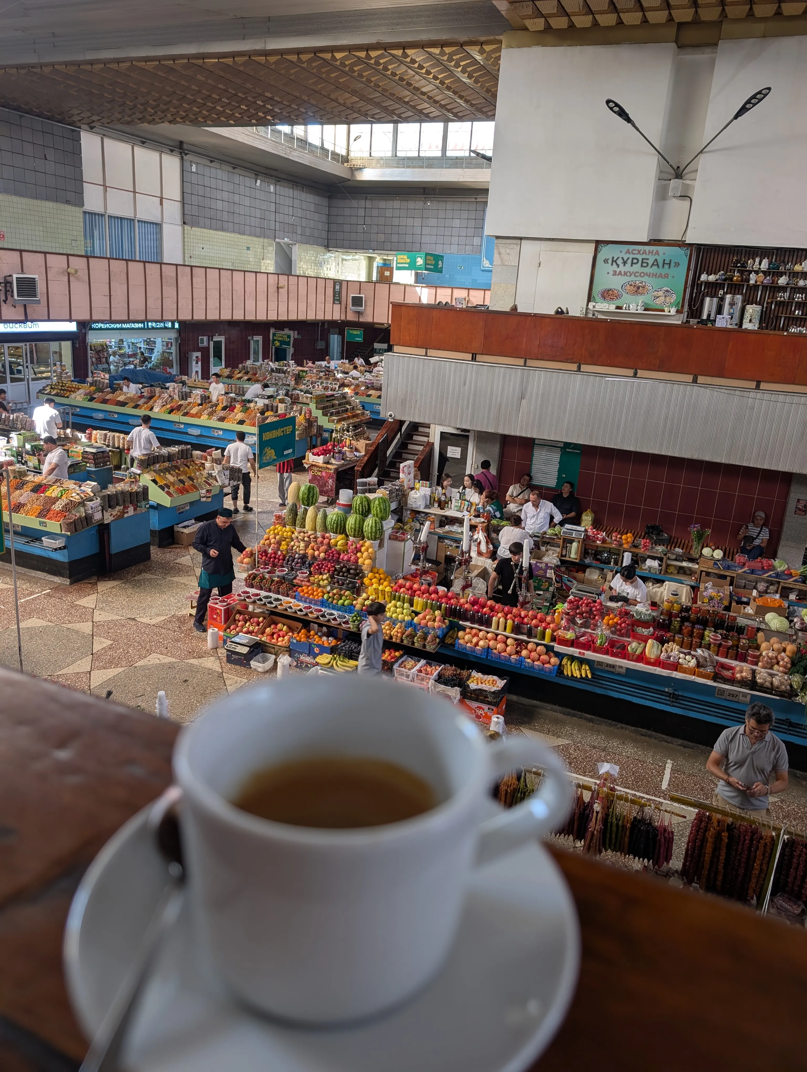 A cup of tea on the wooden rail of Bowler's gallery, with the Green Bazaar spread out below: rows of fruit and spice stalls, traders and shoppers, and strings of churchkhela hanging in the foreground