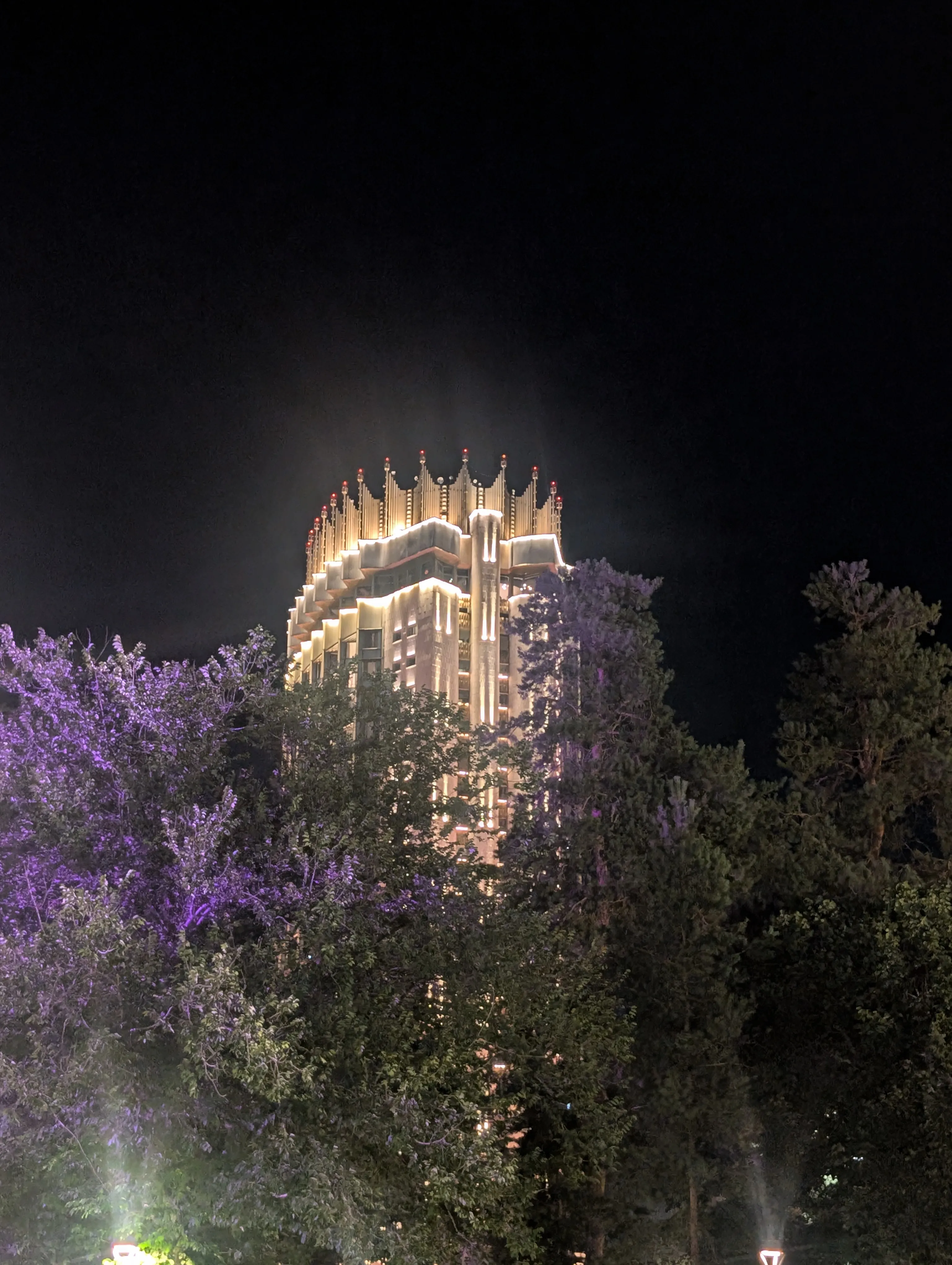 The tower after dark from the end of a tree-lined avenue, every floor and the whole crown picked out in warm light above the empty street