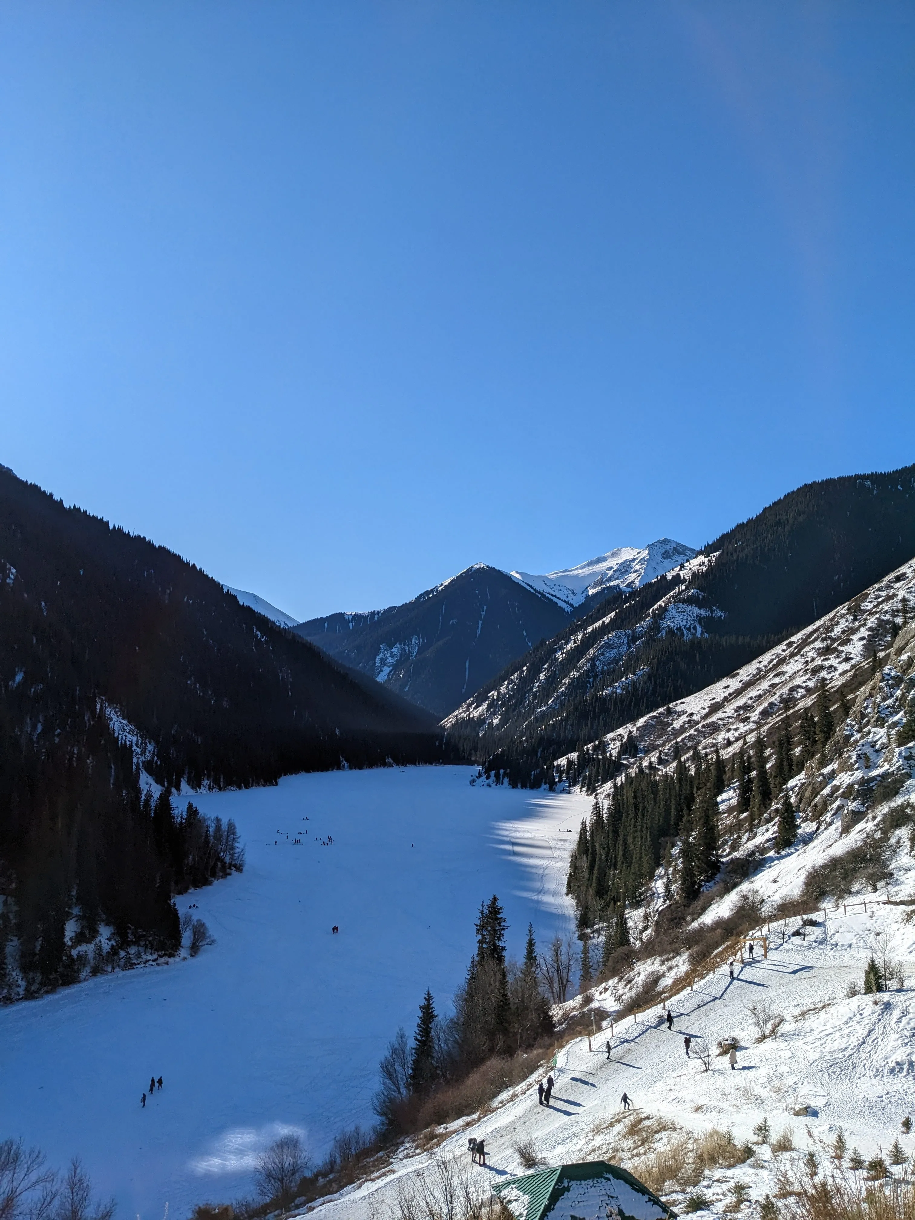 The lower Kolsai lake frozen over in January, seen from the path above it: people walking out across the white ice, dark spruce down both sides of the valley, and snow-covered peaks at its head