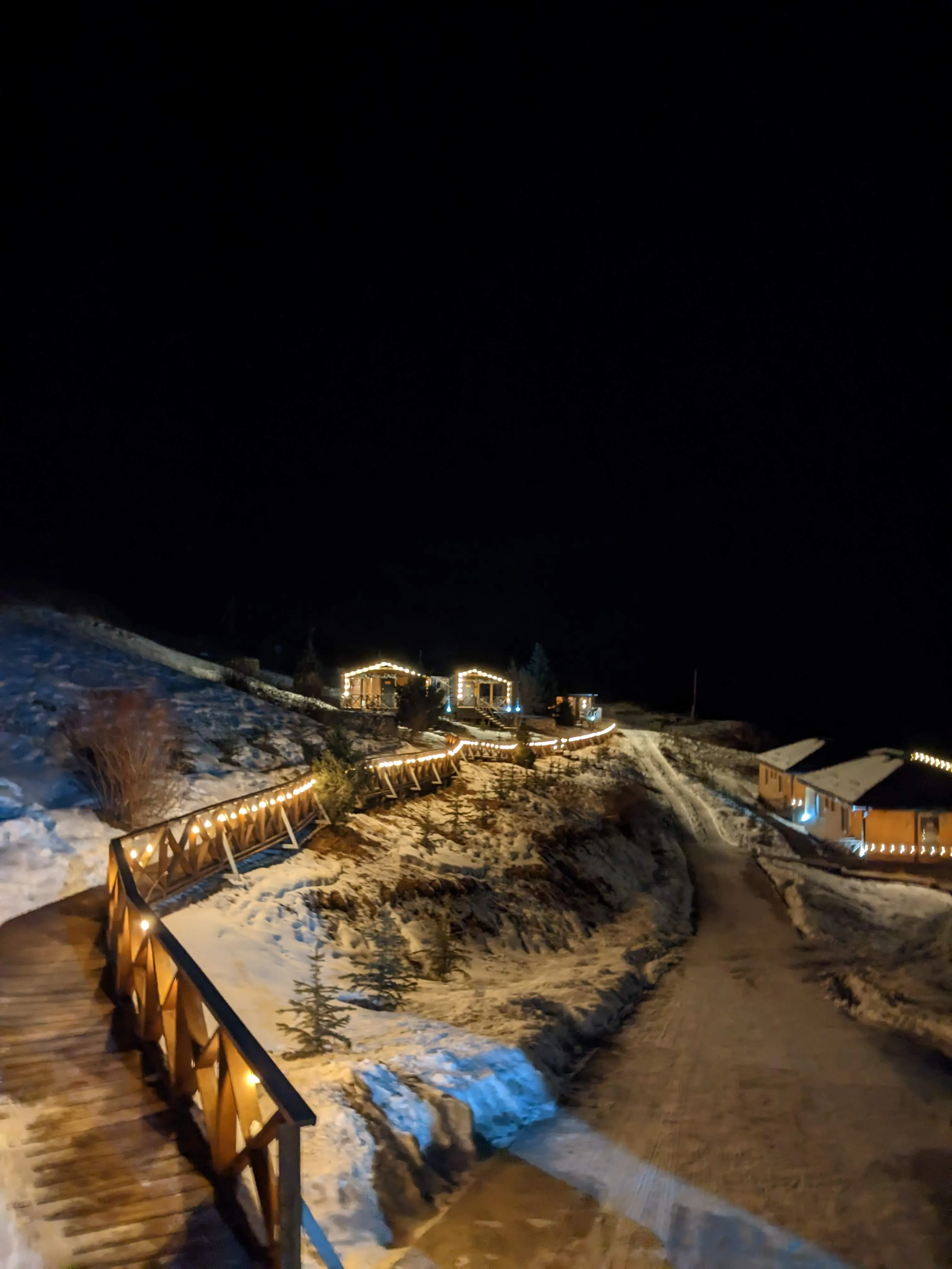 The guesthouse at night in winter: a wooden walkway strung with lights curving up the snowy slope towards lit cabins, and a bare dirt track running past them