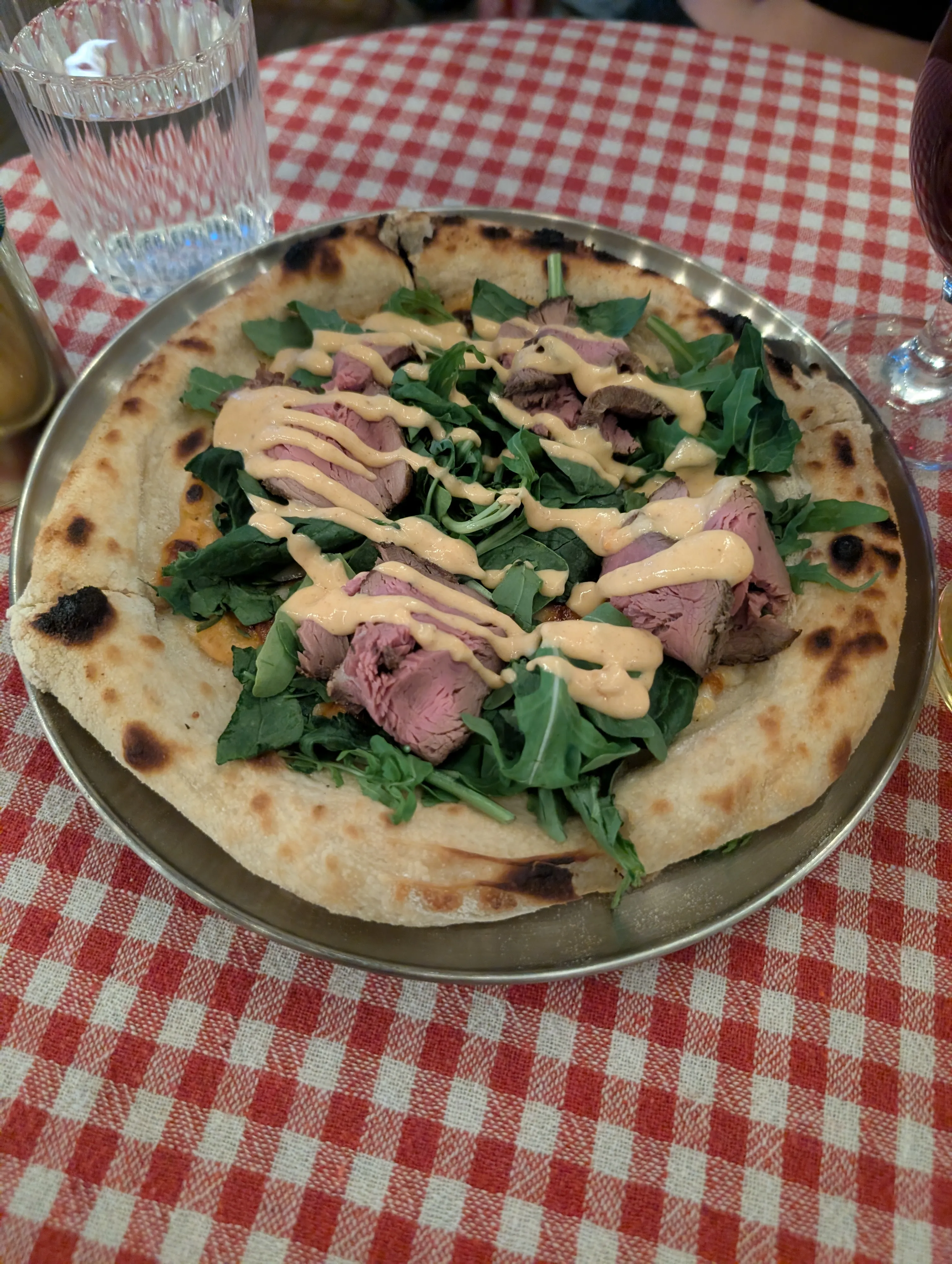 A round pizza with rare roast beef, rocket and a drizzle of aioli on a steel tray over a red gingham cloth at Komok
