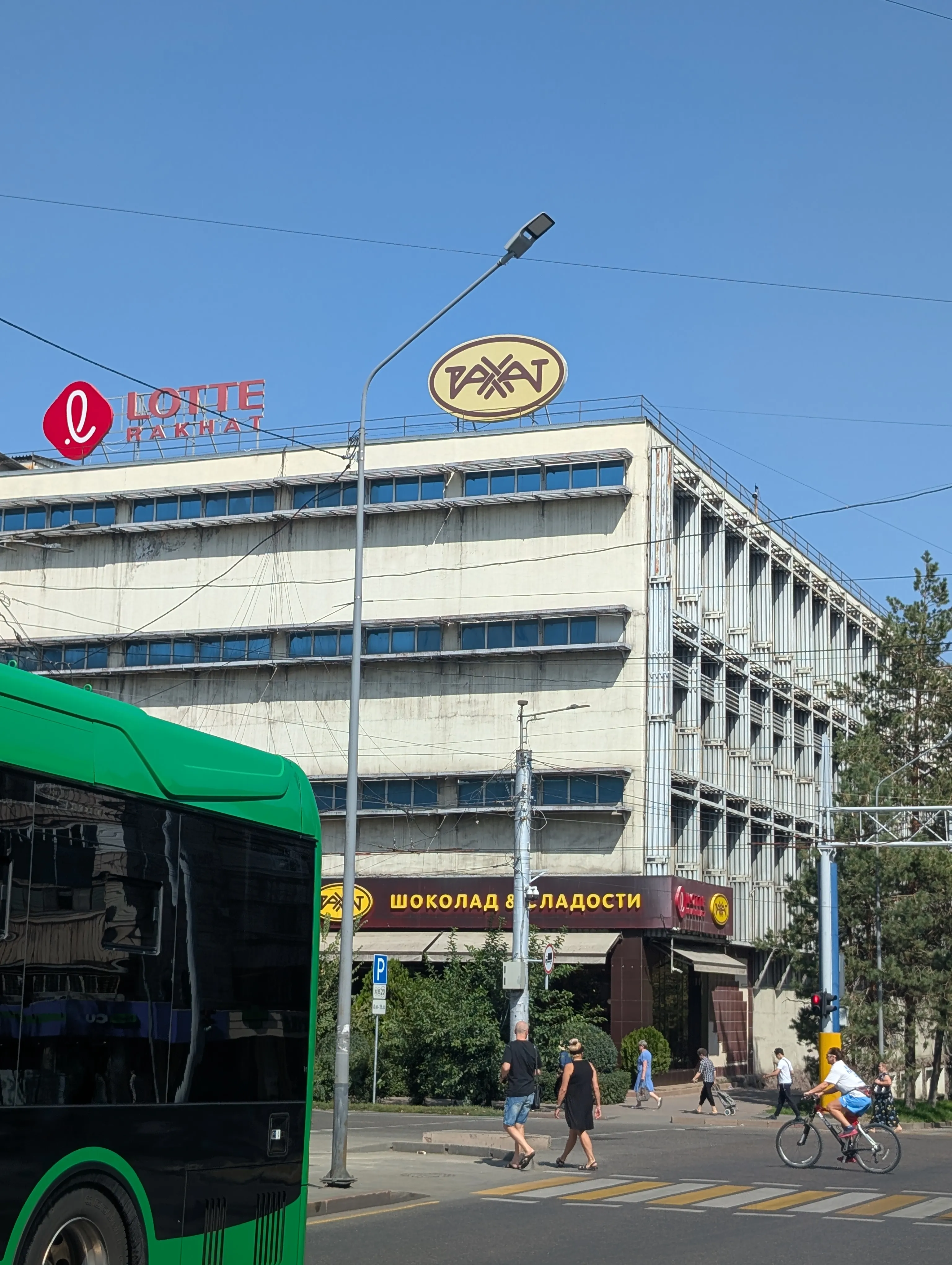 The Rahat factory on Zenkov Street: a long concrete plant with ribbon windows, the yellow Rahat oval and a red LOTTE sign on the roof, a dark shopfront reading 'шоколад и сладости' at street level and a green city bus passing