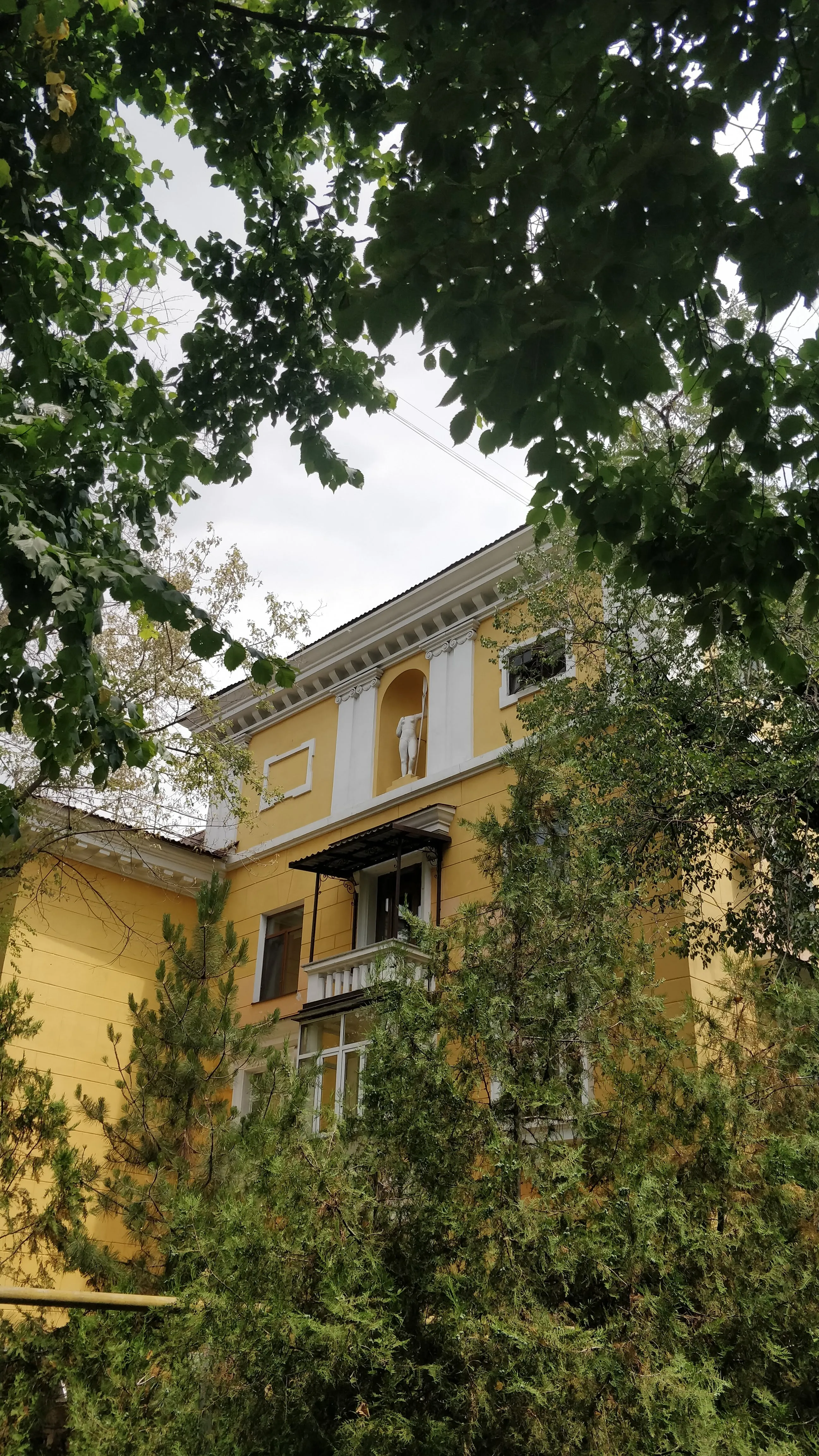 A white statue of a hunter holding a spear, standing in an arched niche high on the yellow facade of an ordinary residential block, framed by trees