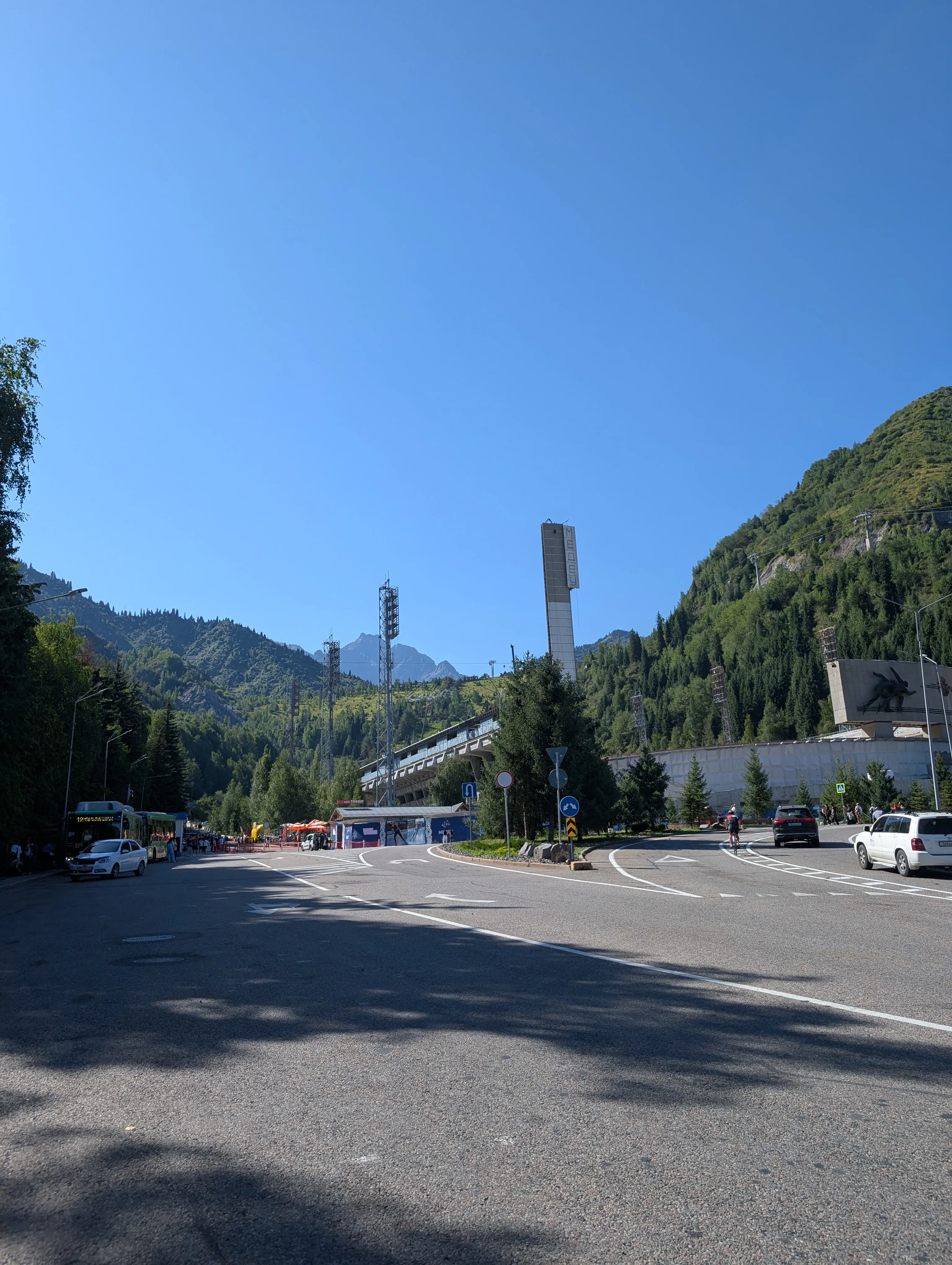 The road and car park at Medeu on a clear morning: buses and taxis waiting, the Medeu tower above the rink, and the mountains behind