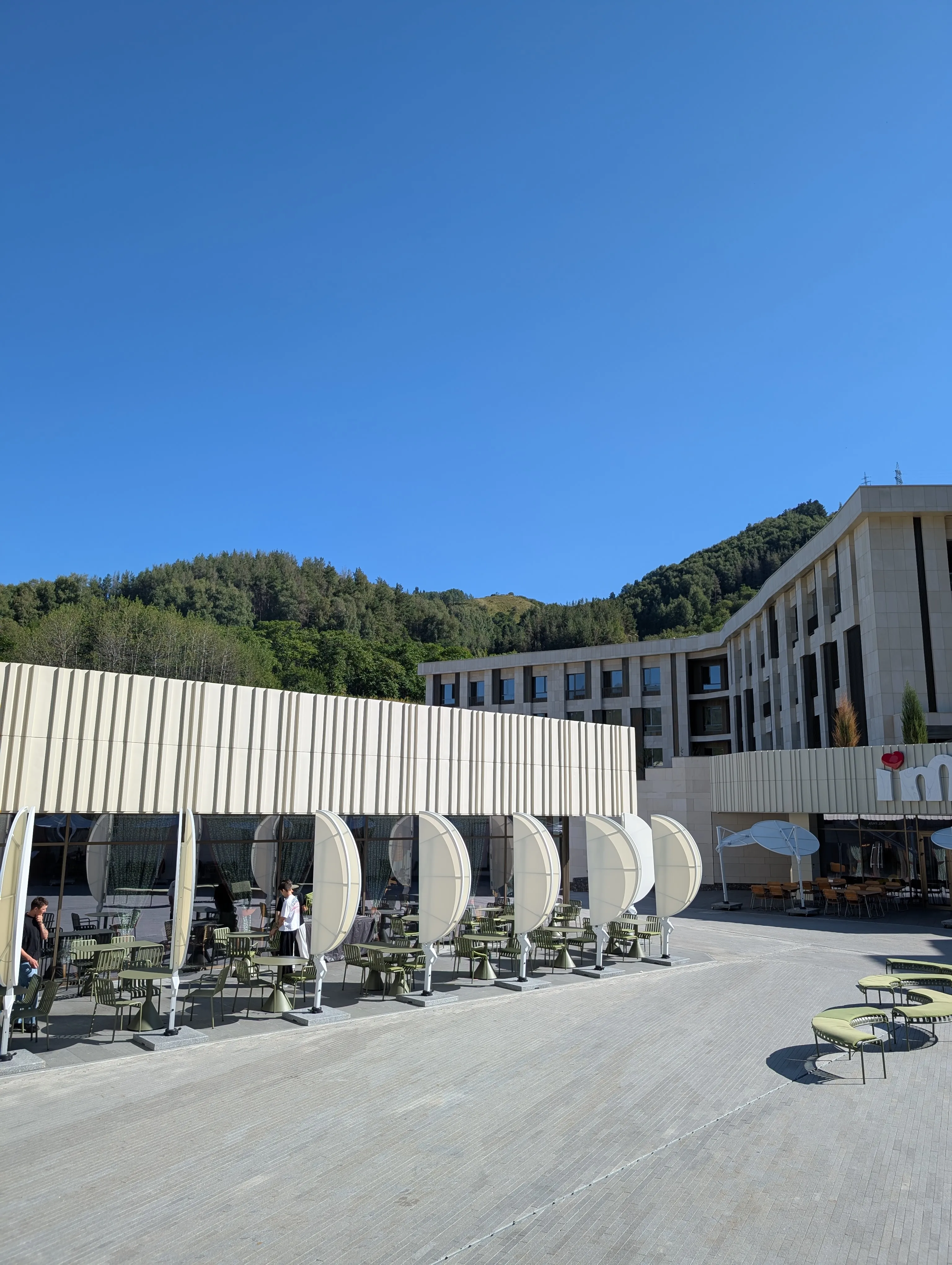 The Del Papa terrace at Medeu: green chairs under shell-shaped parasols on a wide stone plaza, with the wooded ridge behind