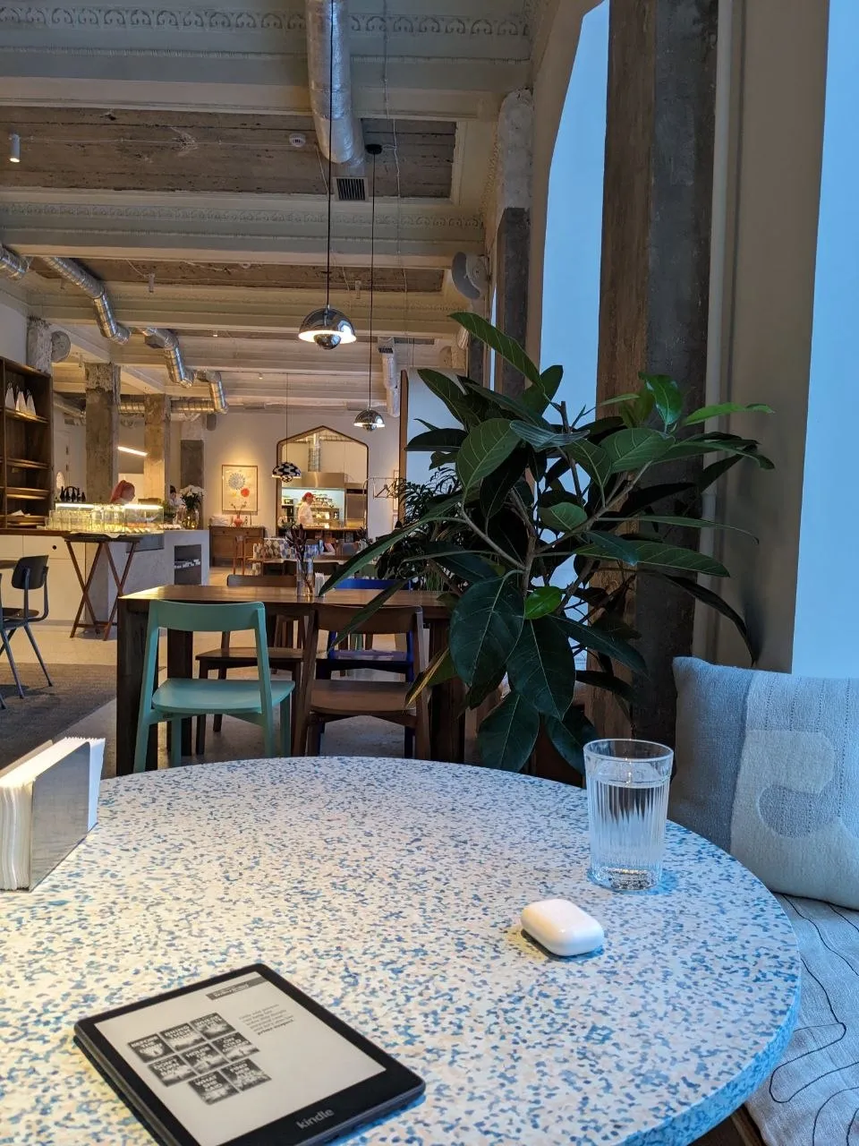 A morning table at Fika: a Kindle and a fluted glass of water on round blue terrazzo, a rubber plant beside the grey banquette, and the bar and long wooden tables under the coffered concrete ceiling behind