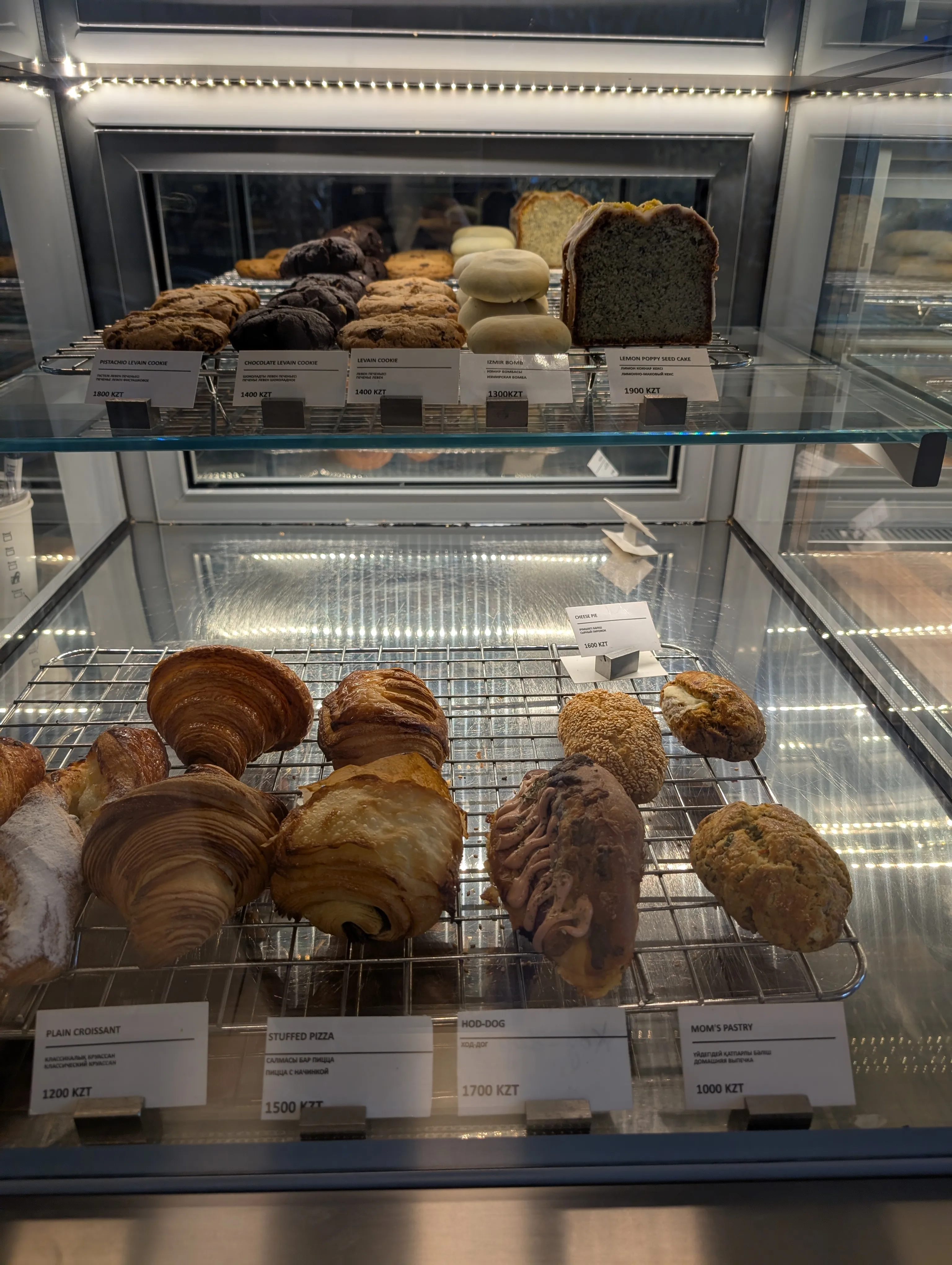 The pastry case at Espressolab: croissants, a chocolate danish and filled pastries on a wire rack with price cards, and levain cookies and a lemon poppy seed cake on the lit shelf above