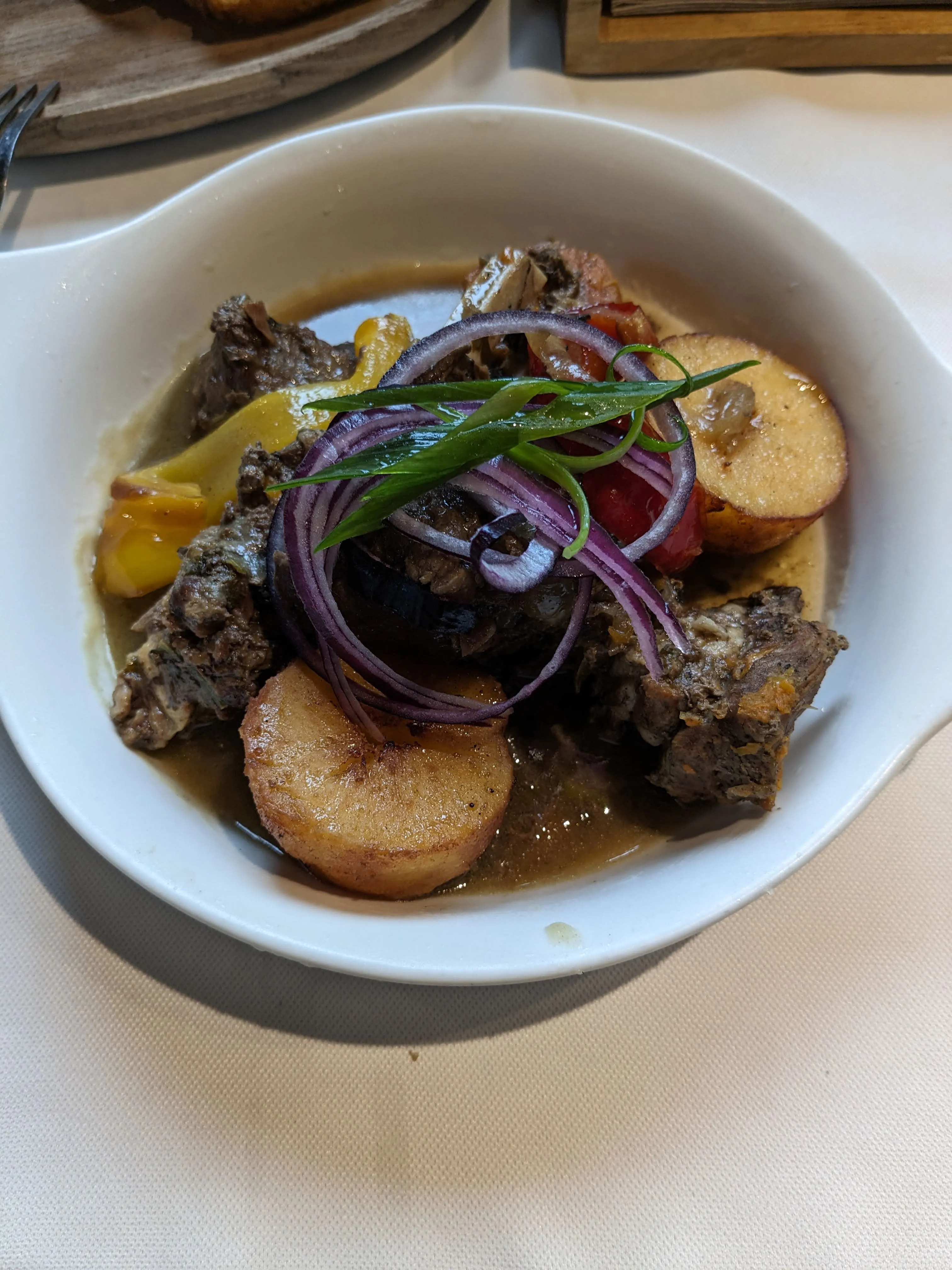 A white bowl of kuyrdak at Qaimaq: dark braised meat and offal in gravy with halved fried potatoes, roast pepper, rings of red onion and a sprig of tarragon over the top