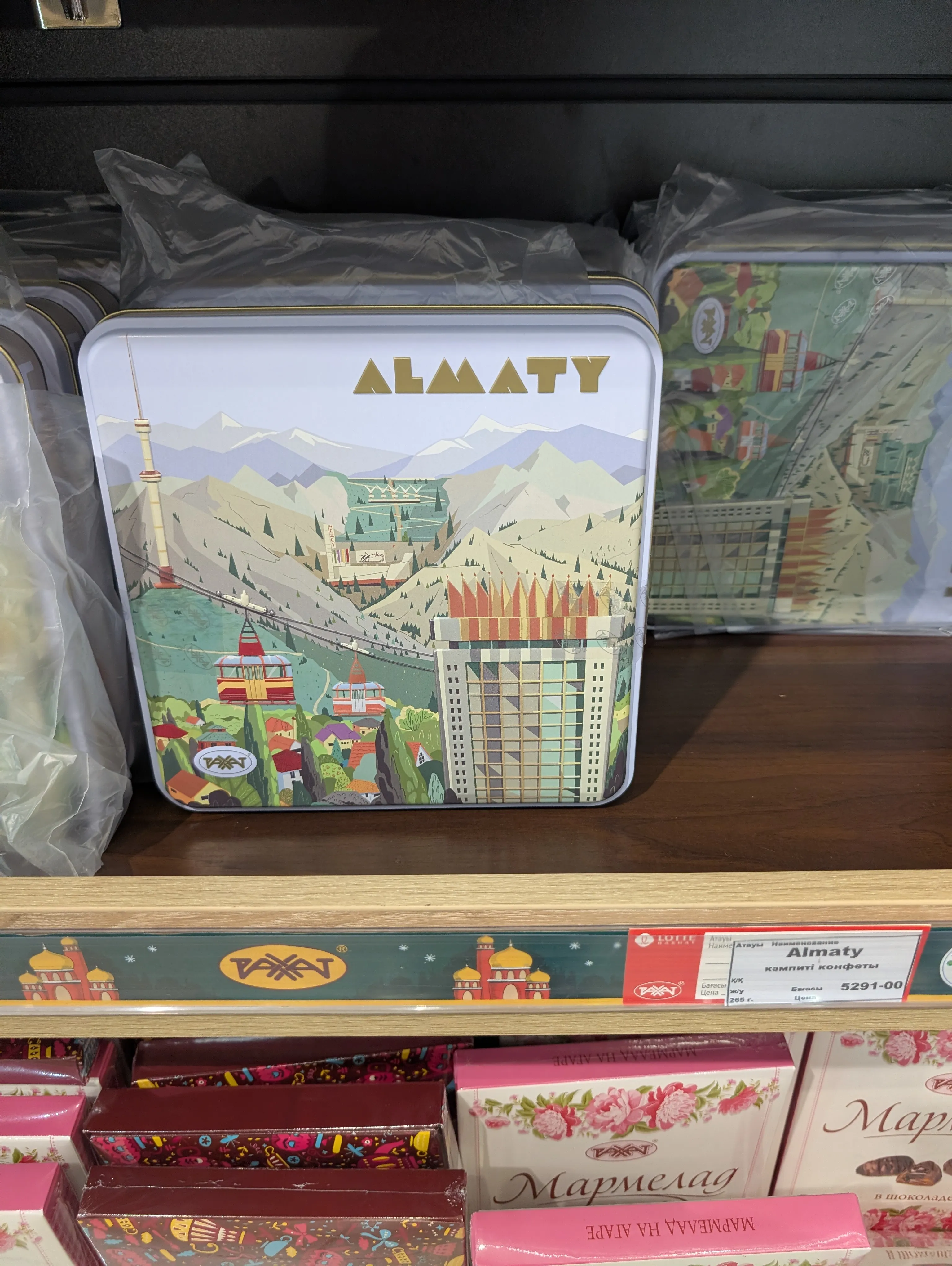 A square Almaty souvenir tin on the shelf: an illustrated city with the television tower, cable car cabins, mountains and the crowned tower of Hotel Kazakhstan, with a tag reading 'Almaty конфеты, 265 г' at 5291 tenge