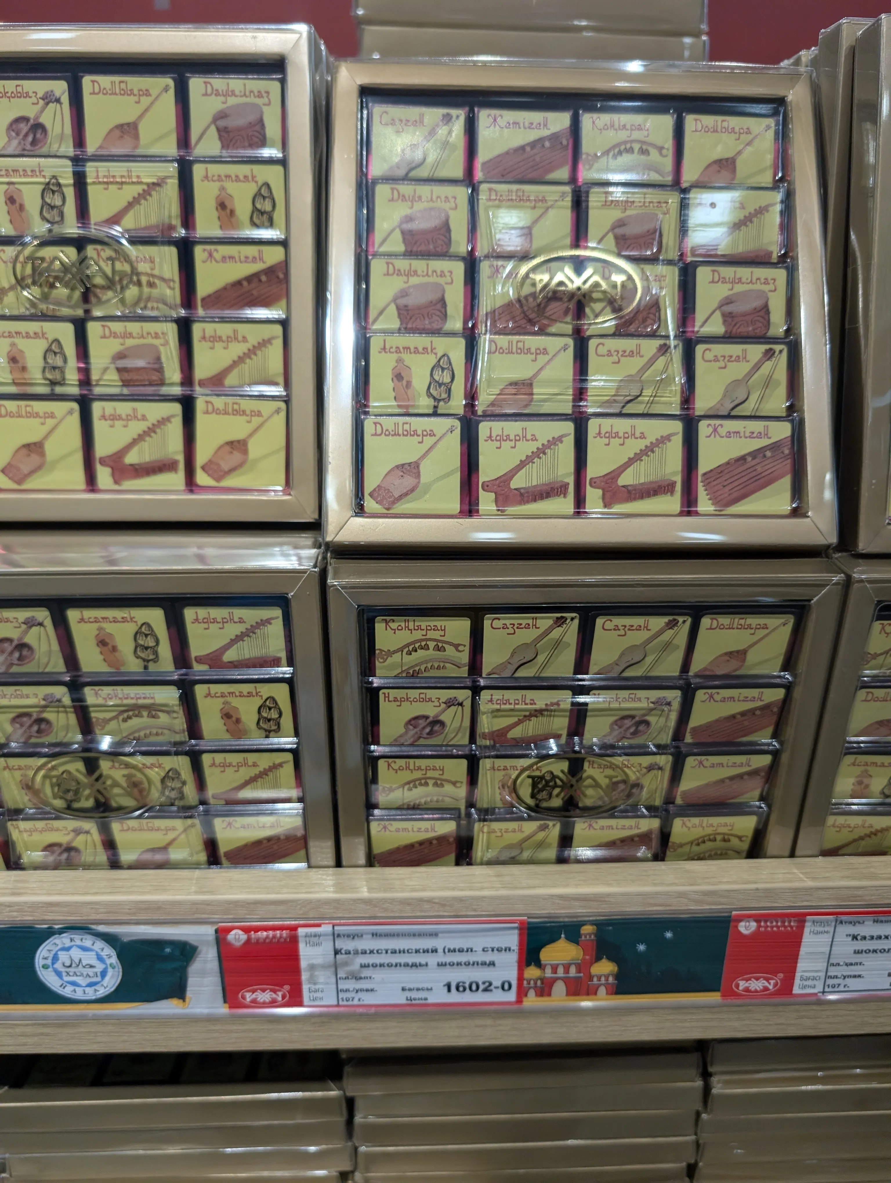 Stacked flat boxes of chocolate, each square printed with a Kazakh instrument and its name — dombyra, kobyz, zhetigen, sazsyrnai — above a shelf tag reading 'Казахстанский' at 1602 tenge