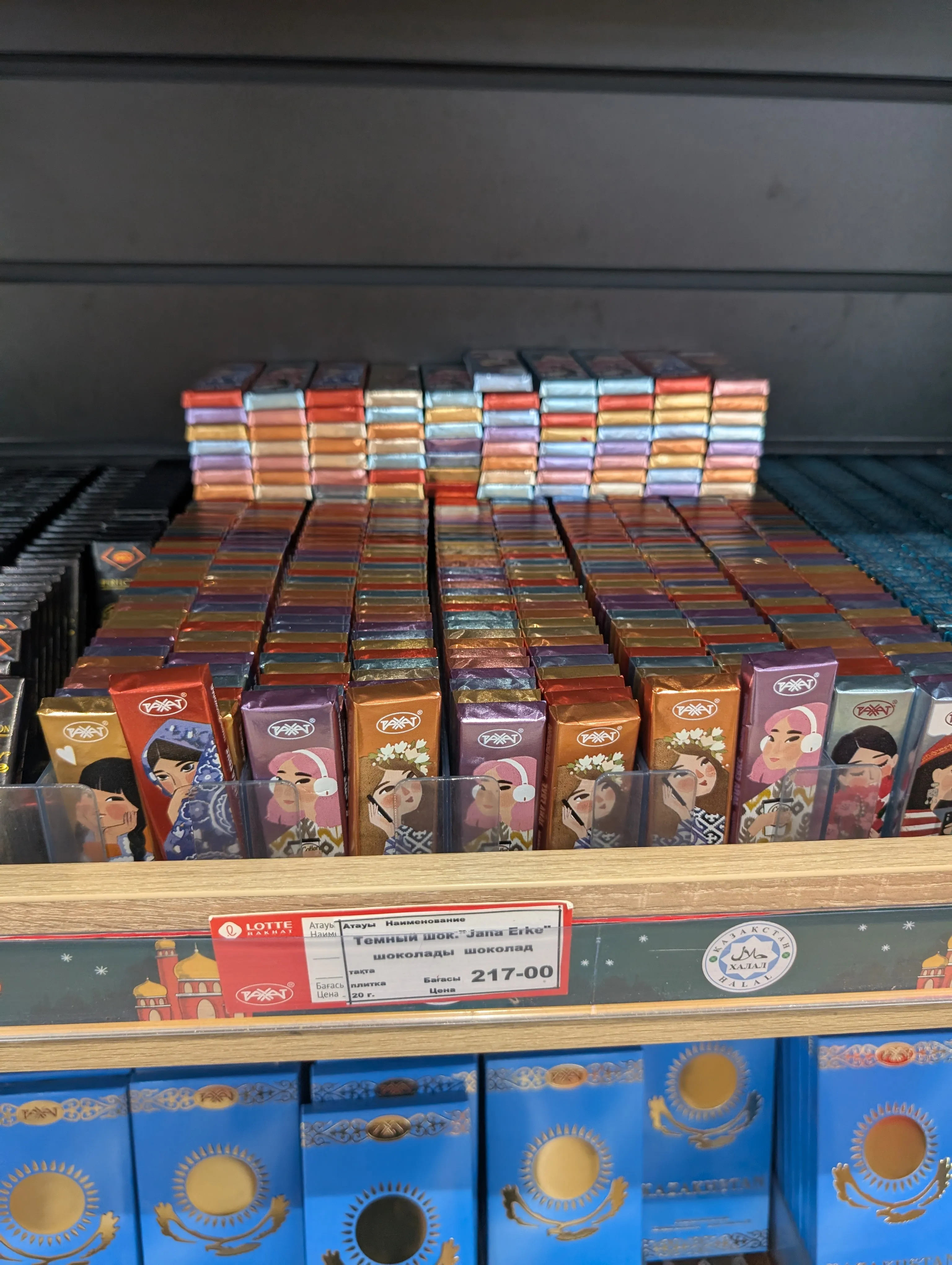 A shelf of small Rahat bars in coloured foil, the fronts illustrated with Kazakh couples in national dress, above a red shelf tag reading 'Темный шок. Jana Erke' at 217 tenge