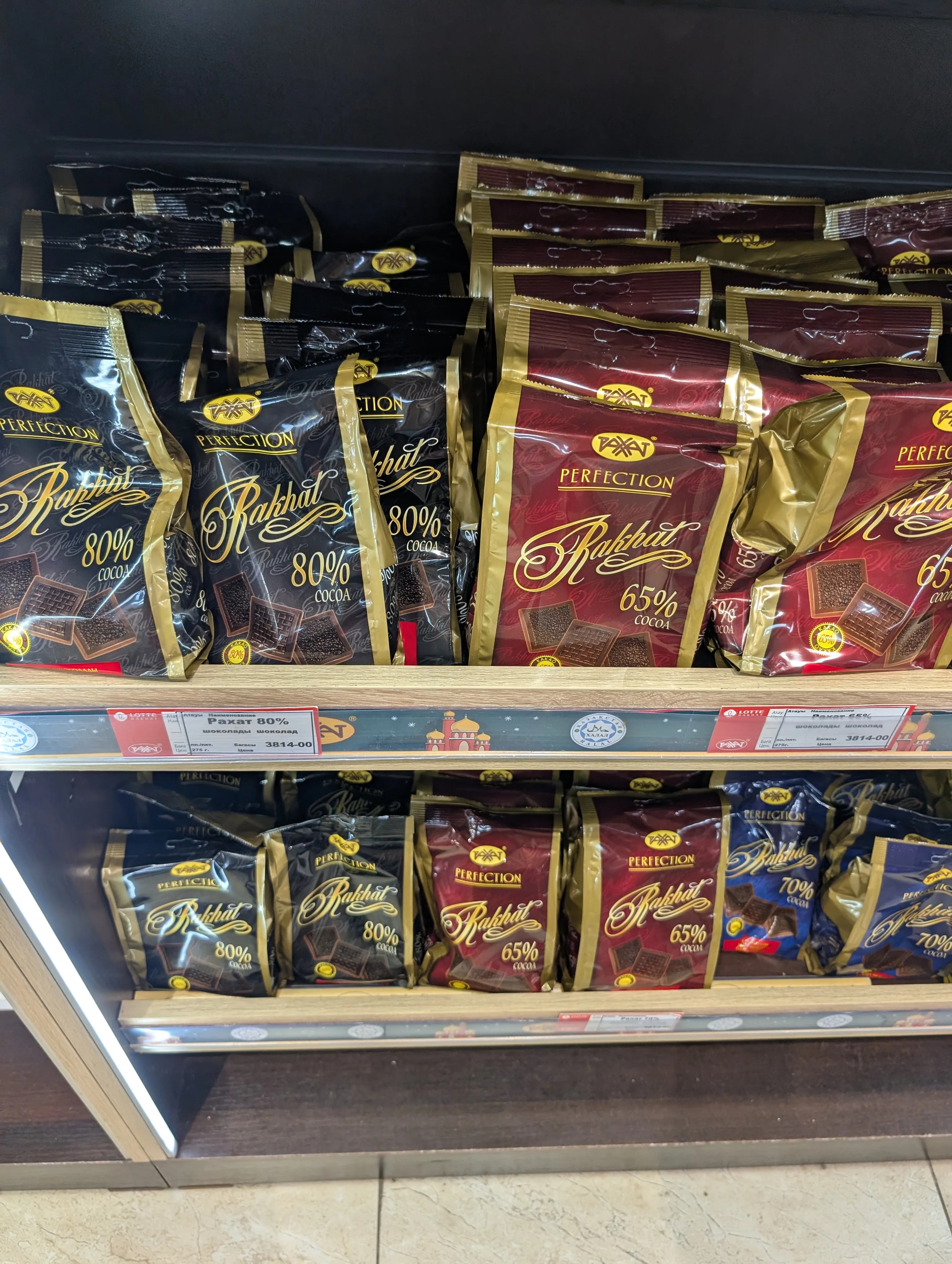 Black and burgundy Rahat Perfection bags marked 80% and 65% cocoa stacked on two shelves, with a shelf tag reading 'Рахат 80%, 275 г' at 3814 tenge