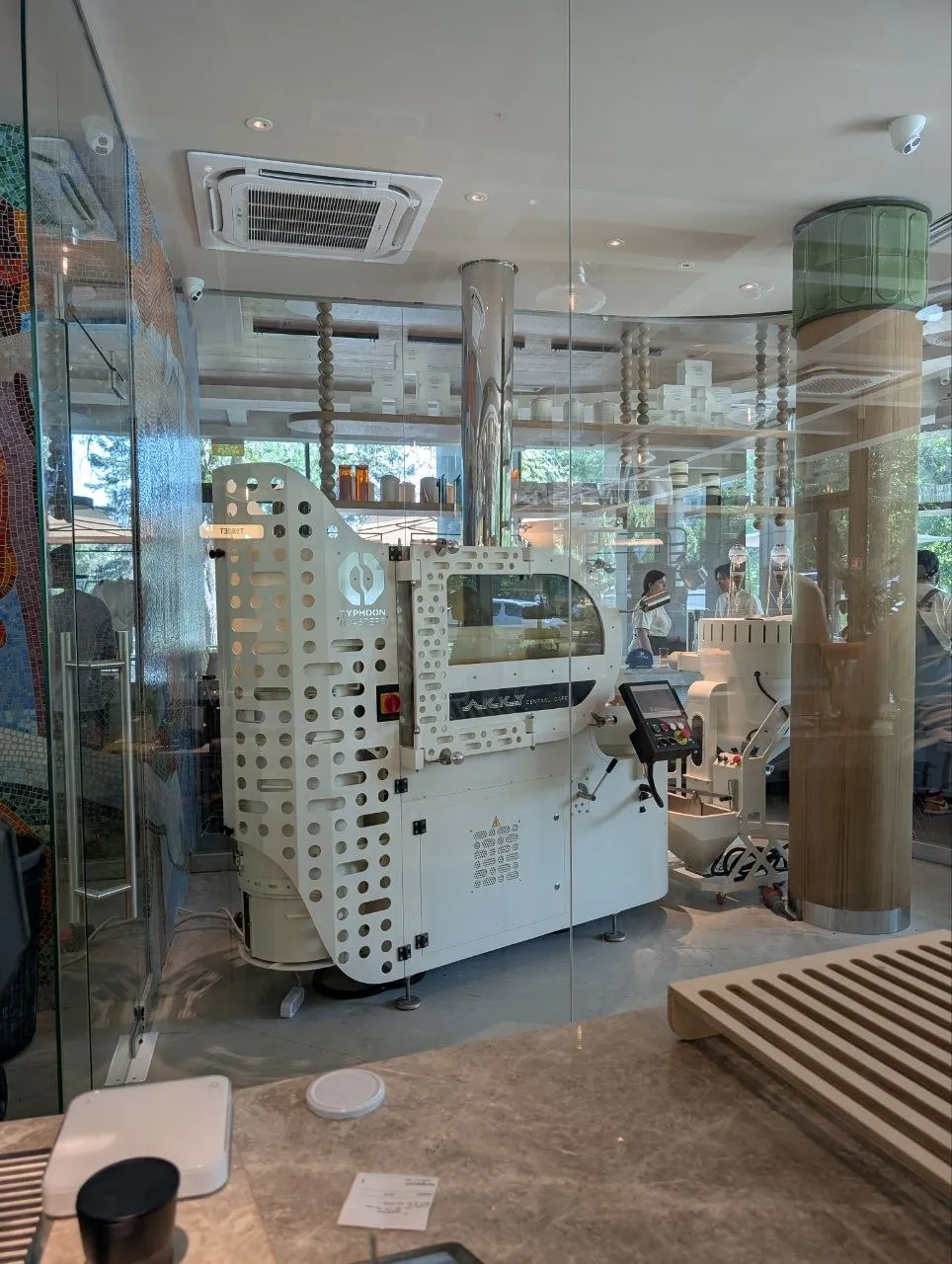 The roastery at Akku behind a glass wall: a white Typhoon Roasters drum machine with a touchscreen panel and a steel chimney to the ceiling, shelves of bean tins and the mosaic wall of the room beyond