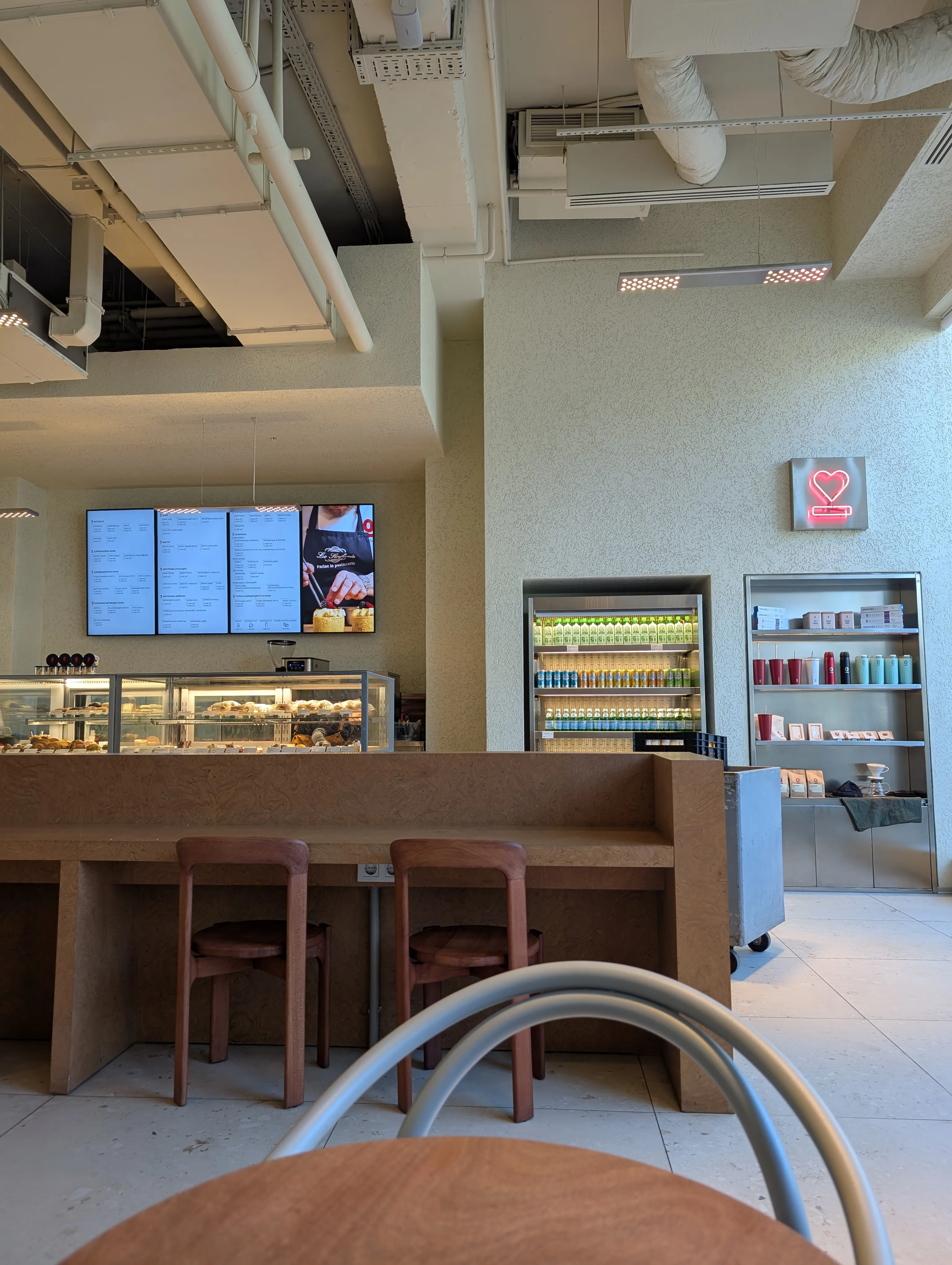 The room at Espressolab in Dostyk Plaza: a pale chipboard bar counter with wooden stools, menu screens above the pastry cases, a drinks fridge and shelves of beans and cups along the far wall