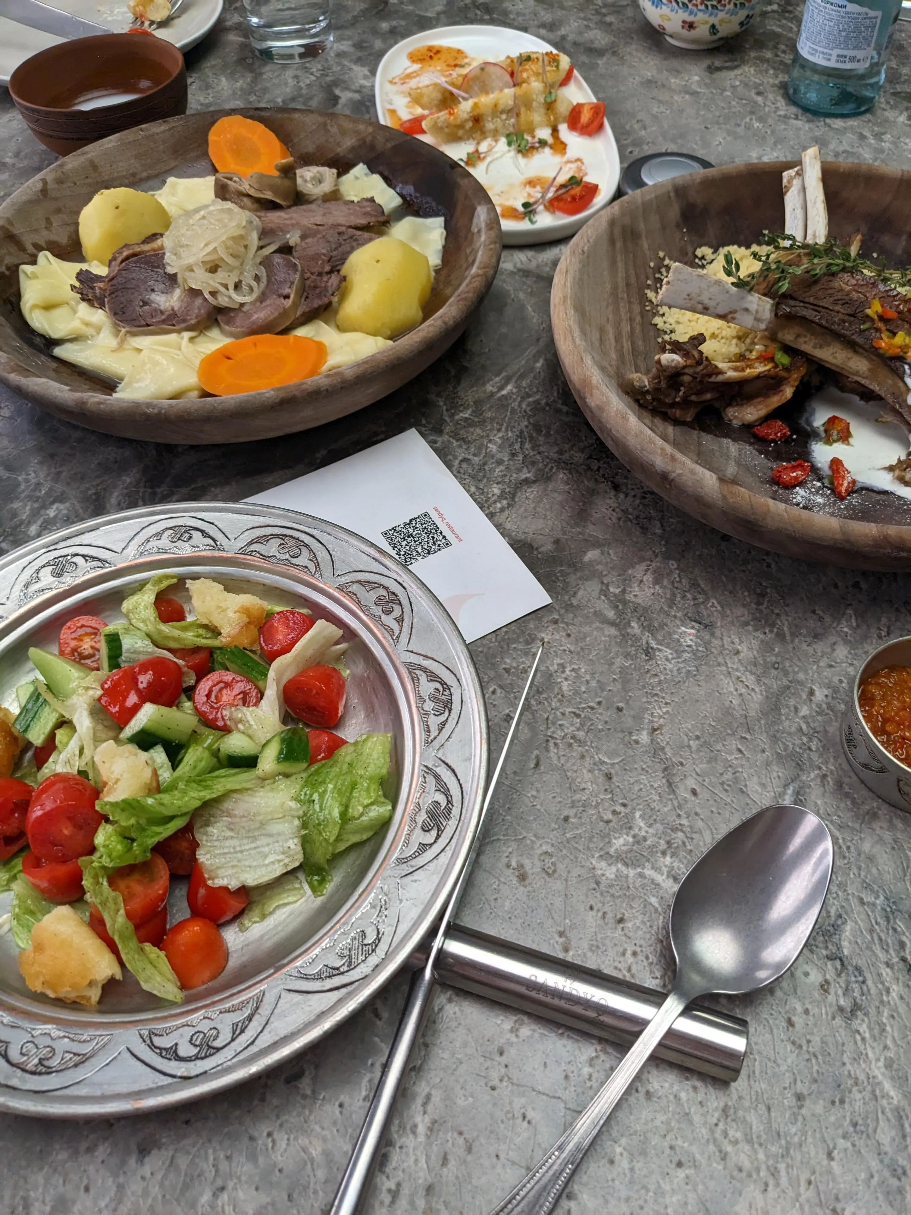 A table at Sandyq: beshbarmak in a wooden bowl with sheets of noodle, boiled meat, potato and rounds of carrot, a rack of lamb in a second wooden bowl, and a tomato and cucumber salad on a patterned metal plate