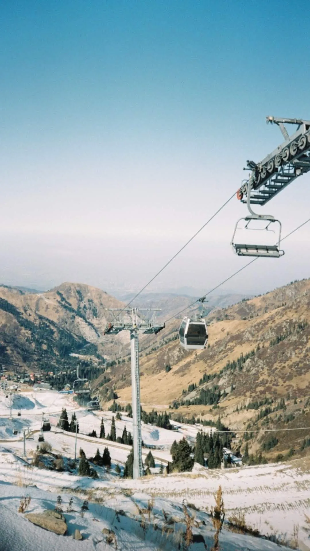 Looking back down the gorge from the Shymbulak lifts: a gondola climbing past its pylon, an empty chairlift overhead, the first snow lying in patches across brown slopes, and the city plain hazy in the distance