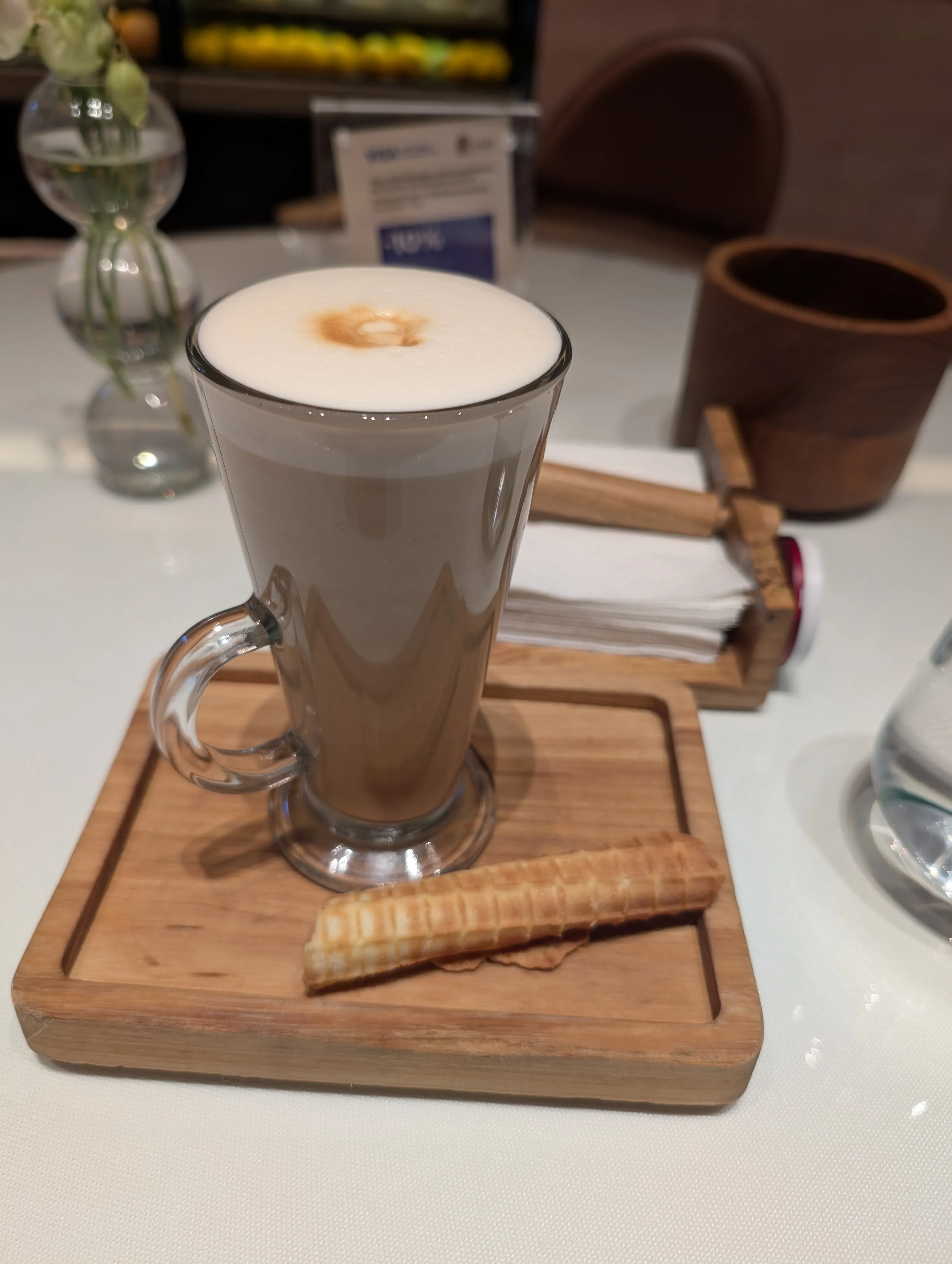An irimshik latte at Tary: a tall glass on a wooden board, dense white foam with a single caramel-coloured spot at its centre, a rolled wafer beside it