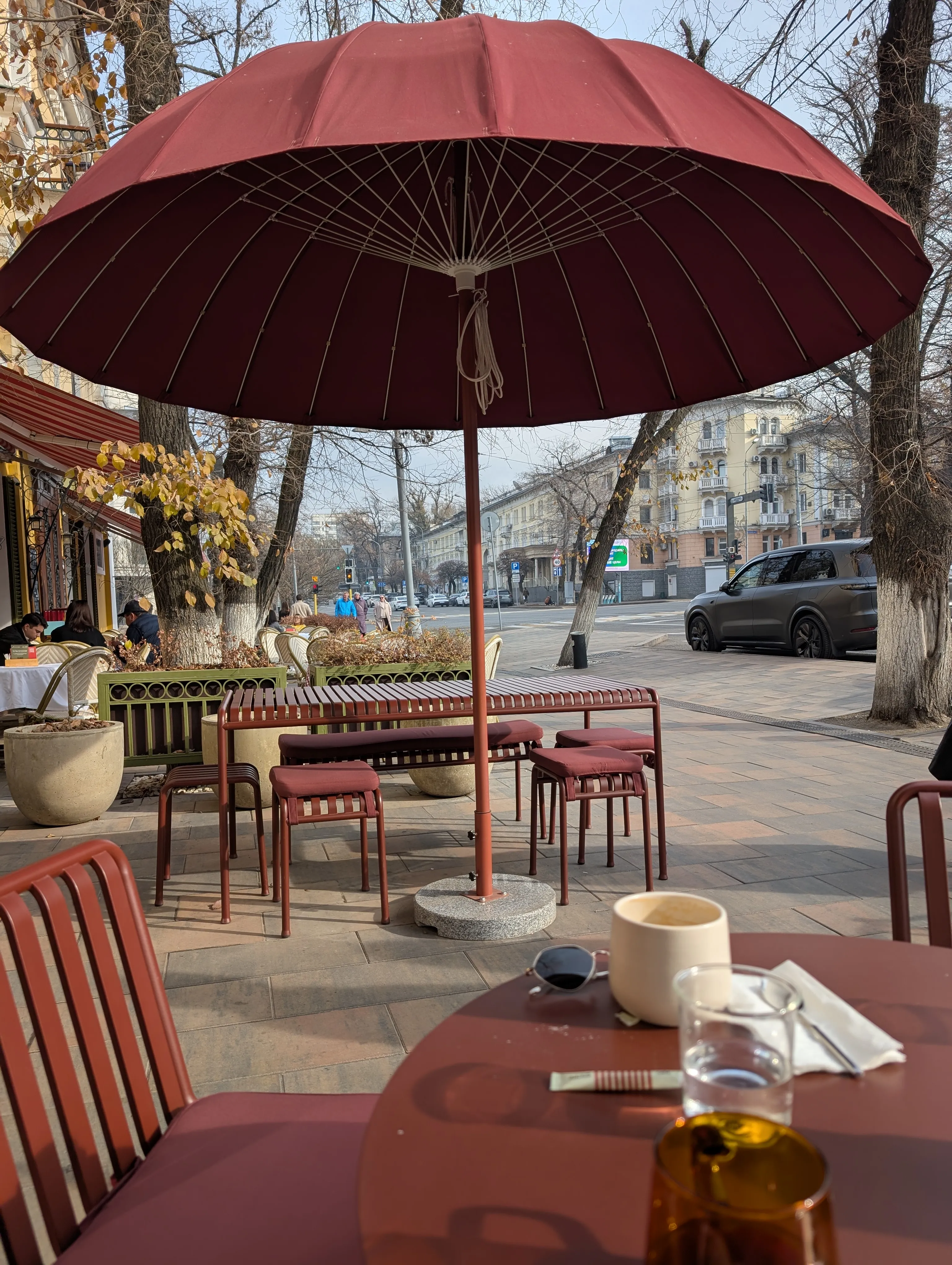 The street terrace at Julius in autumn: a wide dark-red parasol over terracotta tables and stools, a cup of coffee and a glass of water on the table in the foreground, and the yellowing avenue trees beyond