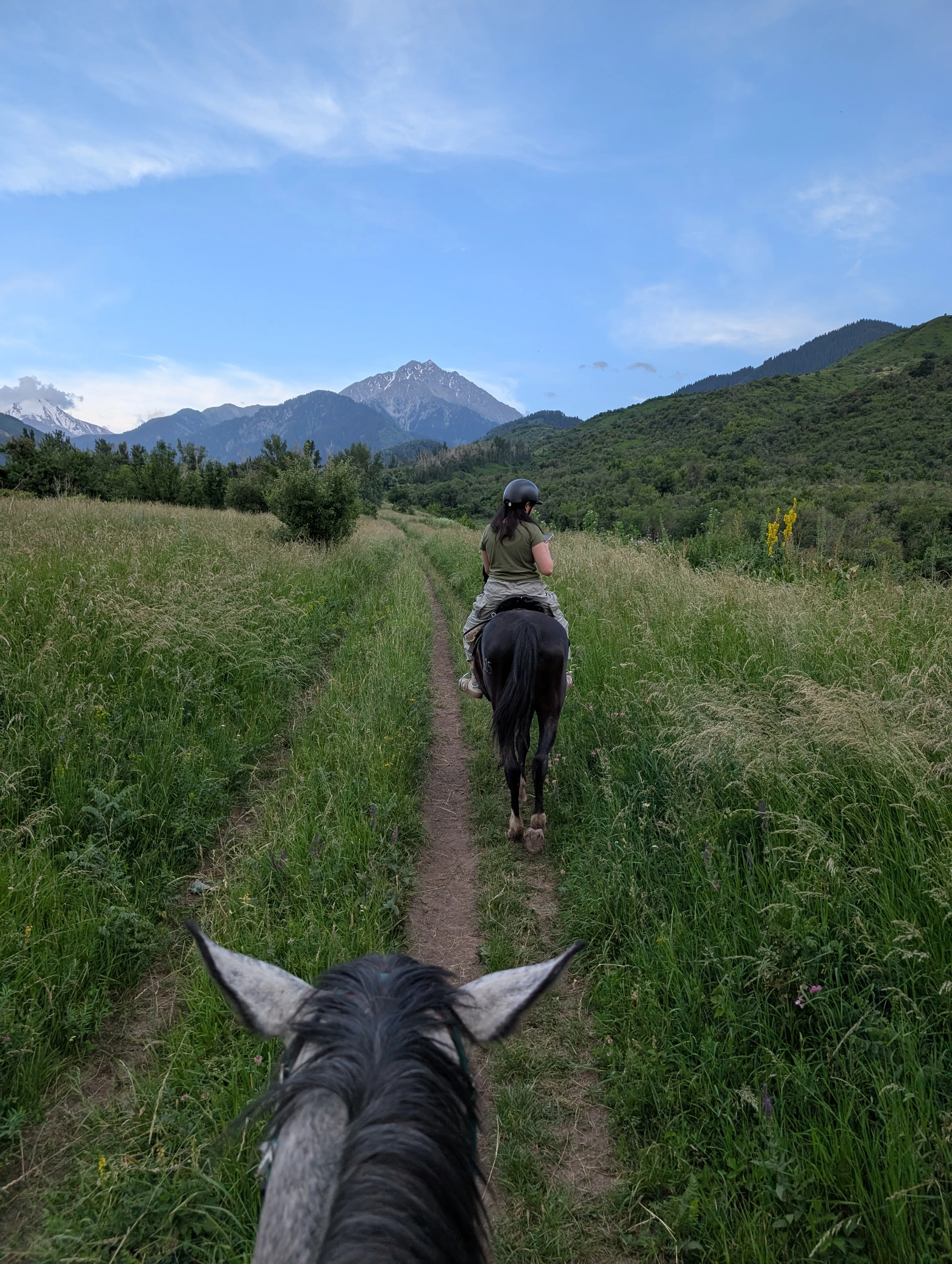Riding a narrow trail through tall summer grass at Almaty Hills, another rider ahead and the mountains beyond