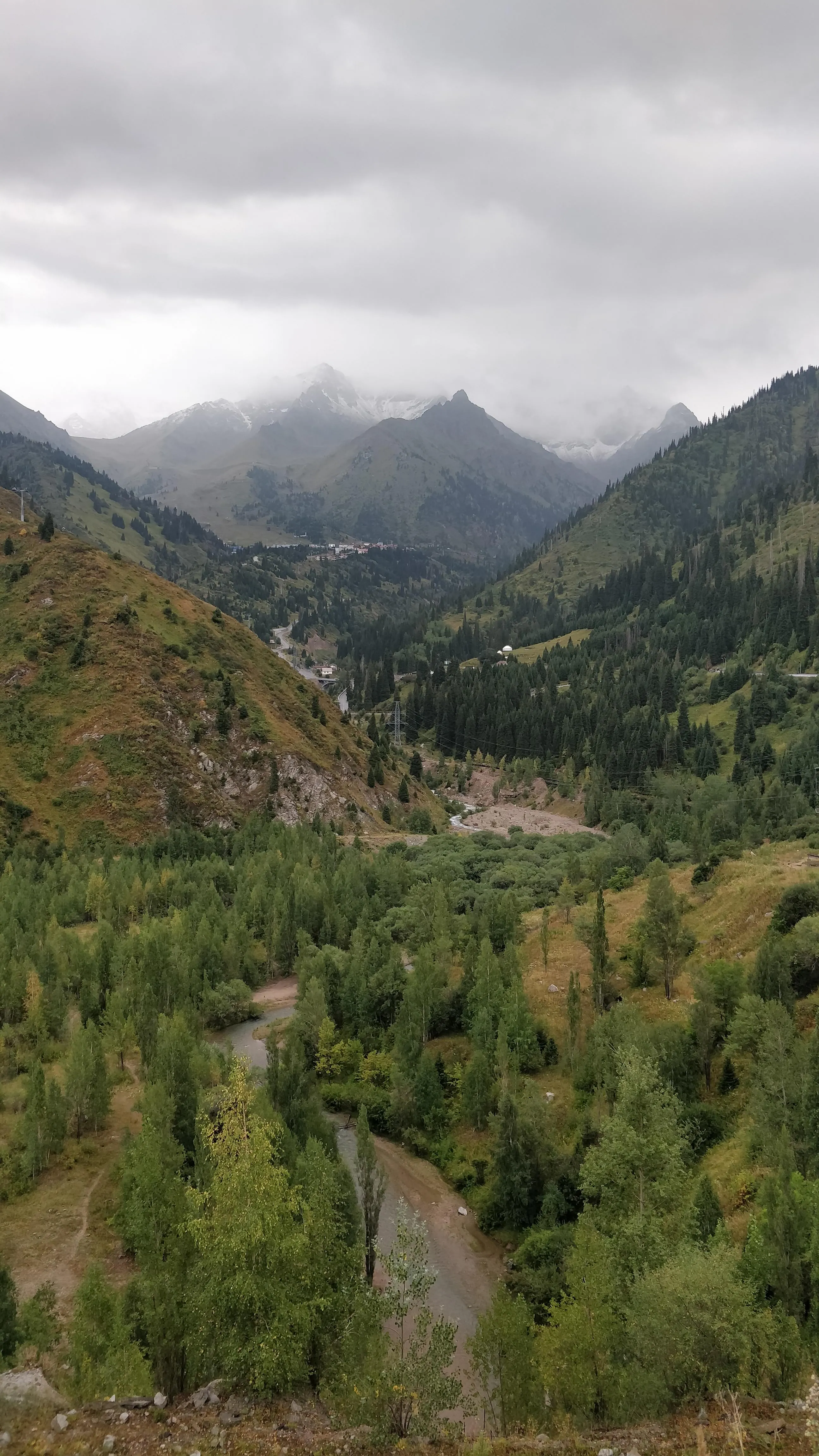 The view from the top of the Medeu steps: the river below, the gorge road climbing towards Shymbulak, and snowy peaks under cloud at the head of the valley