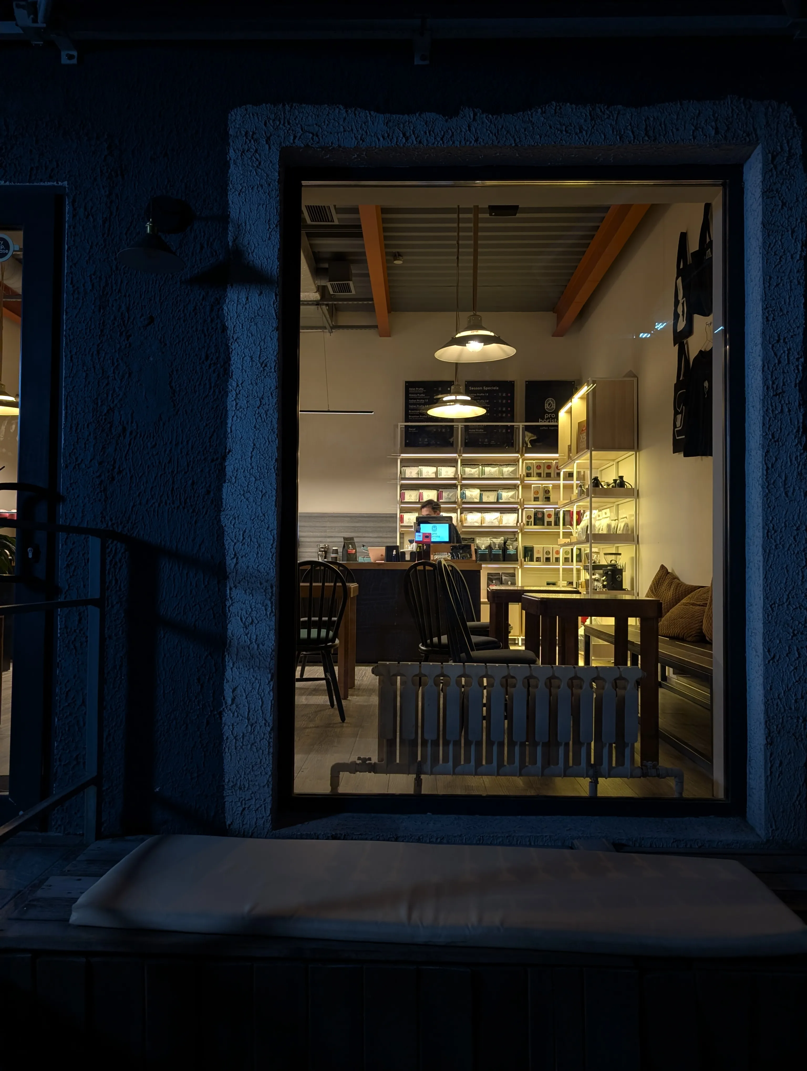 Probarista seen at night through its window: retail shelves of beans and a board listing their roast profiles