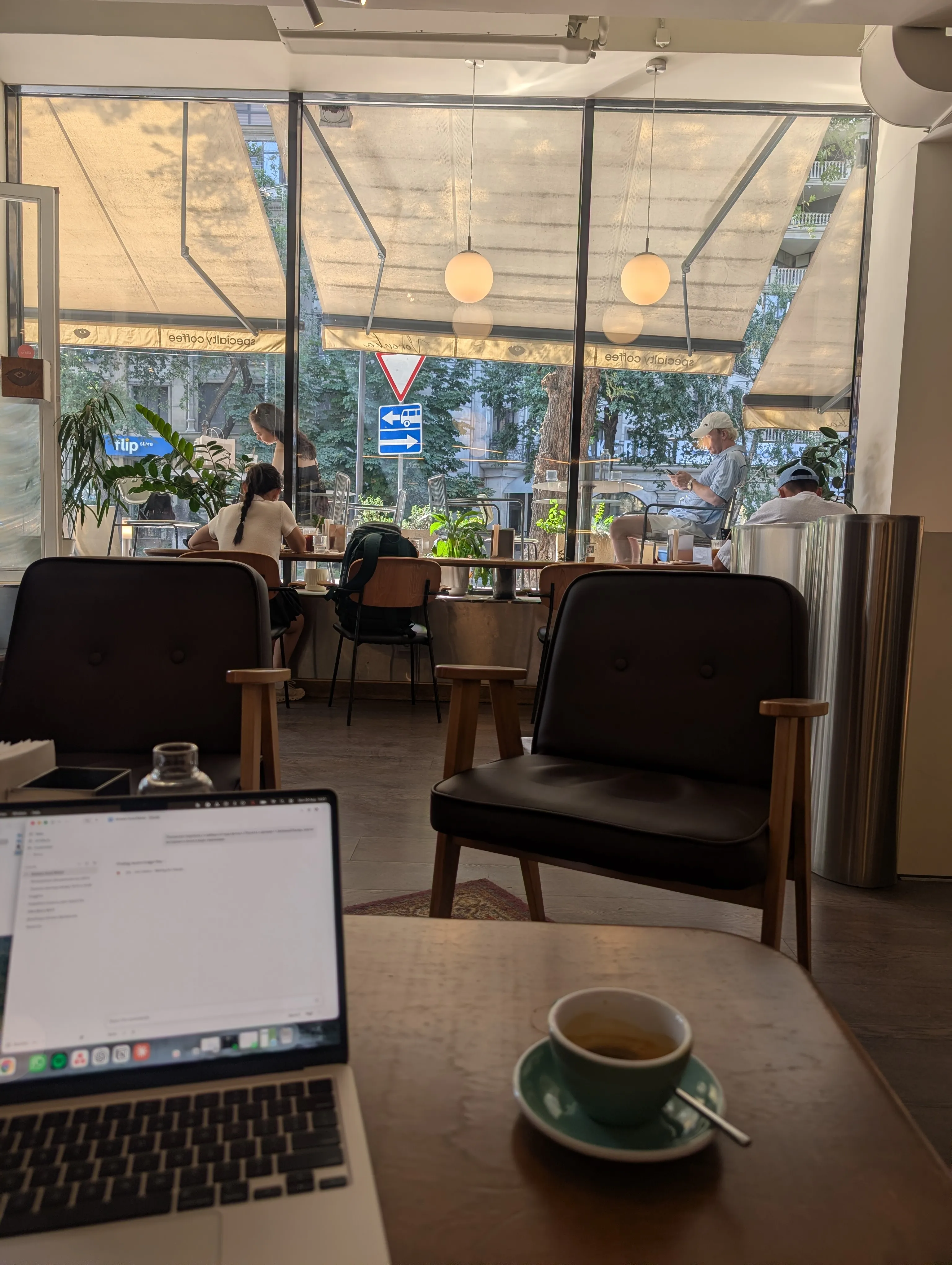 The window room at Voronka from a work table: an open laptop and a green cup on the wood, two dark leather armchairs facing the full-height glass, and the street trees beyond it