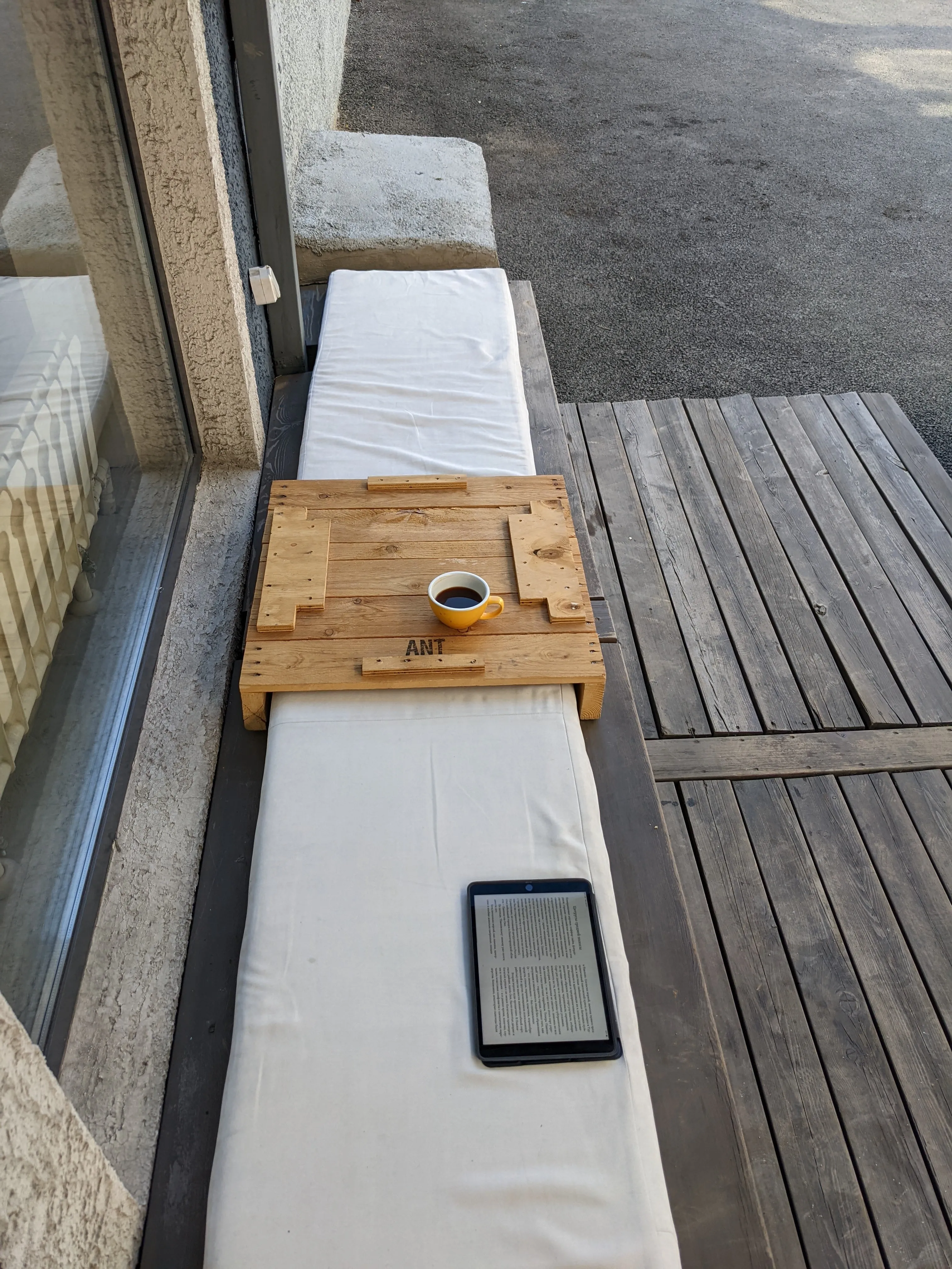 A cup of black coffee on a low pallet table outside Probarista, set on a long bench with pale cushions along the window, an e-reader lying open beside it